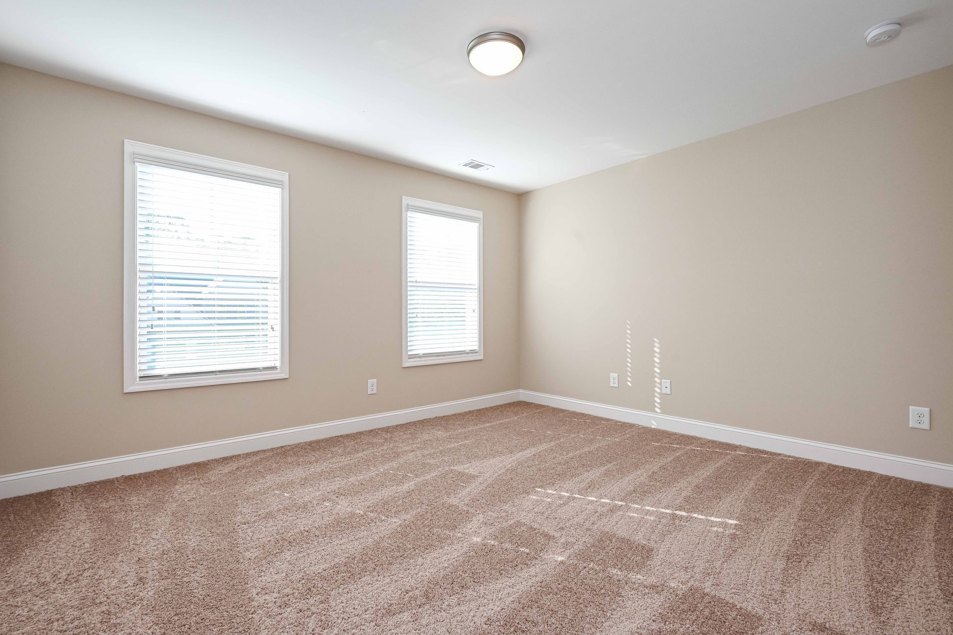 Empty beige carpeted room with two windows and white trim, lit by a ceiling light