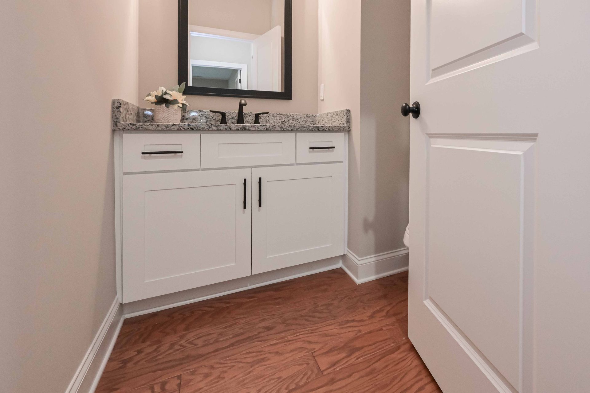 Small hallway bathroom vanity with white cabinets, mirror, and wood floor, seen through an open door