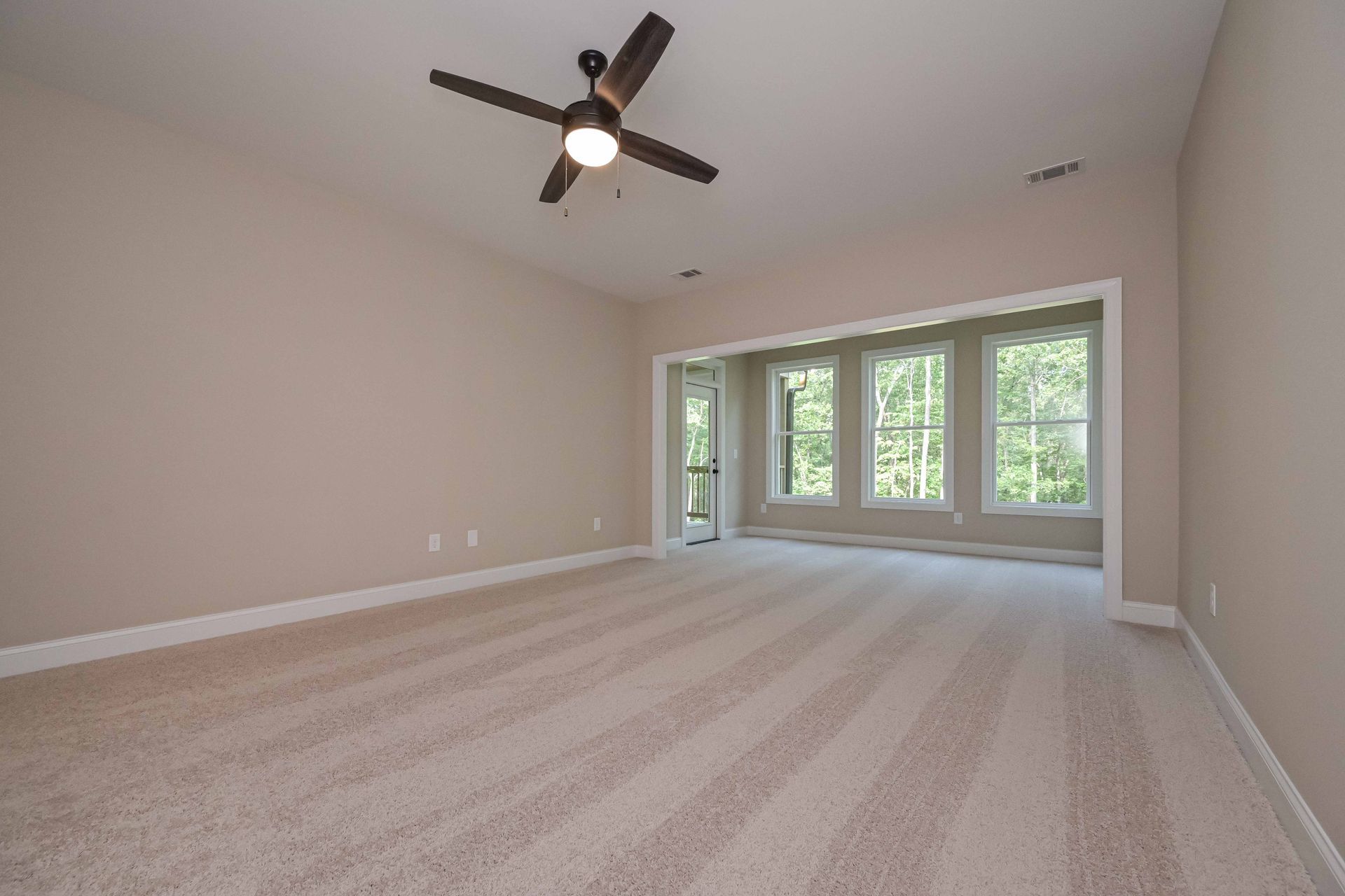 Empty beige living room with ceiling fan, carpet, and large windows overlooking trees
