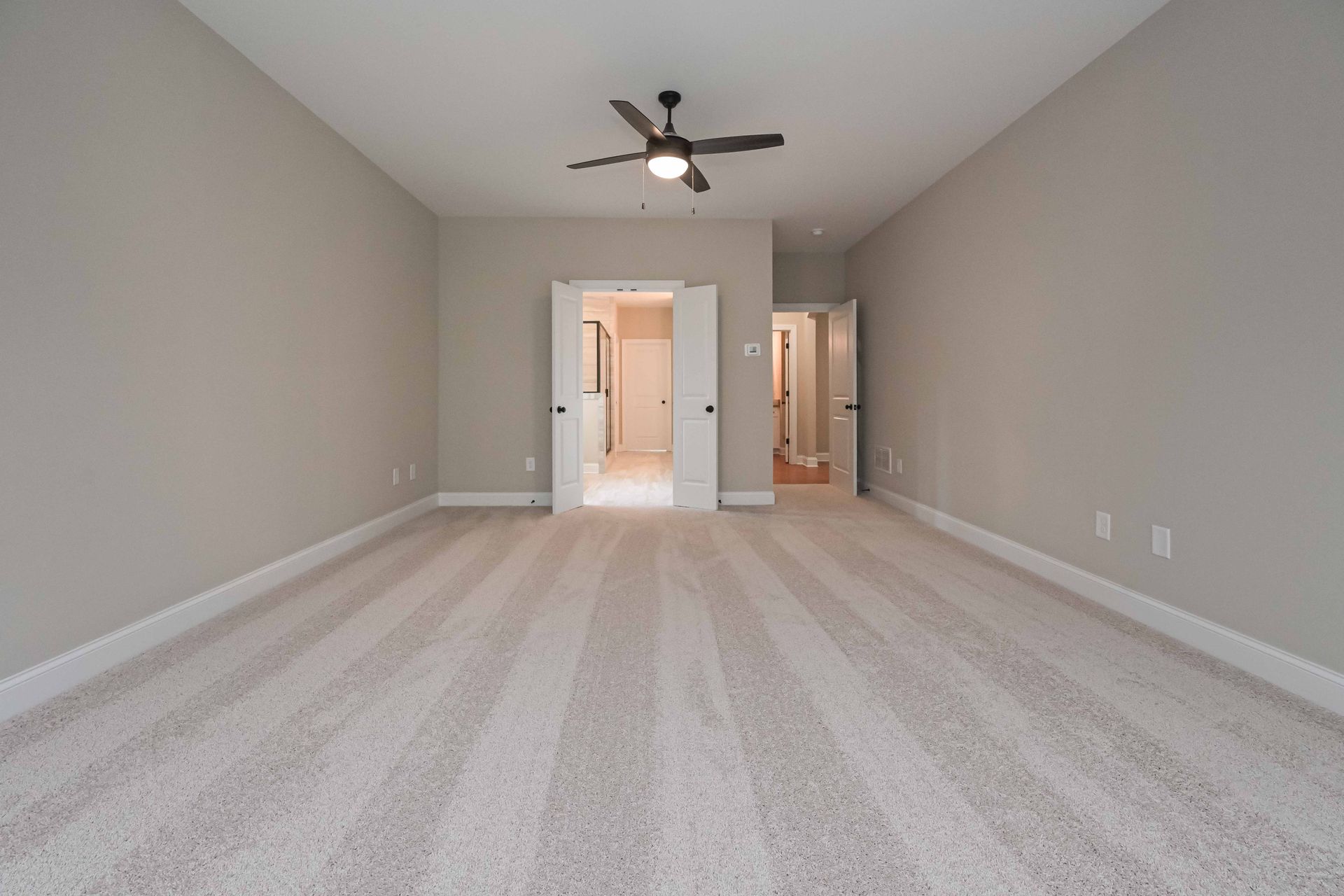 Empty beige carpeted room with gray walls, ceiling fan, and doorway to a bathroom and hallway.
