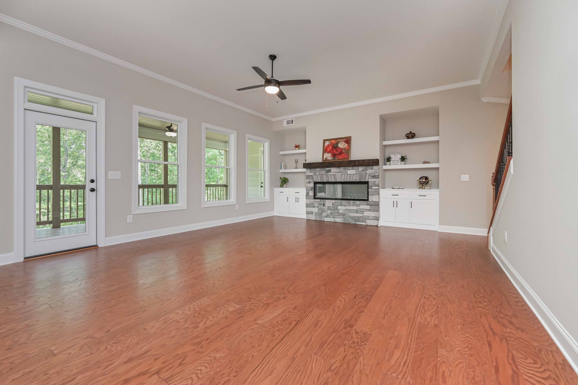 Empty living room with hardwood floors, large windows, ceiling fan, and built-in shelves around a fireplace area