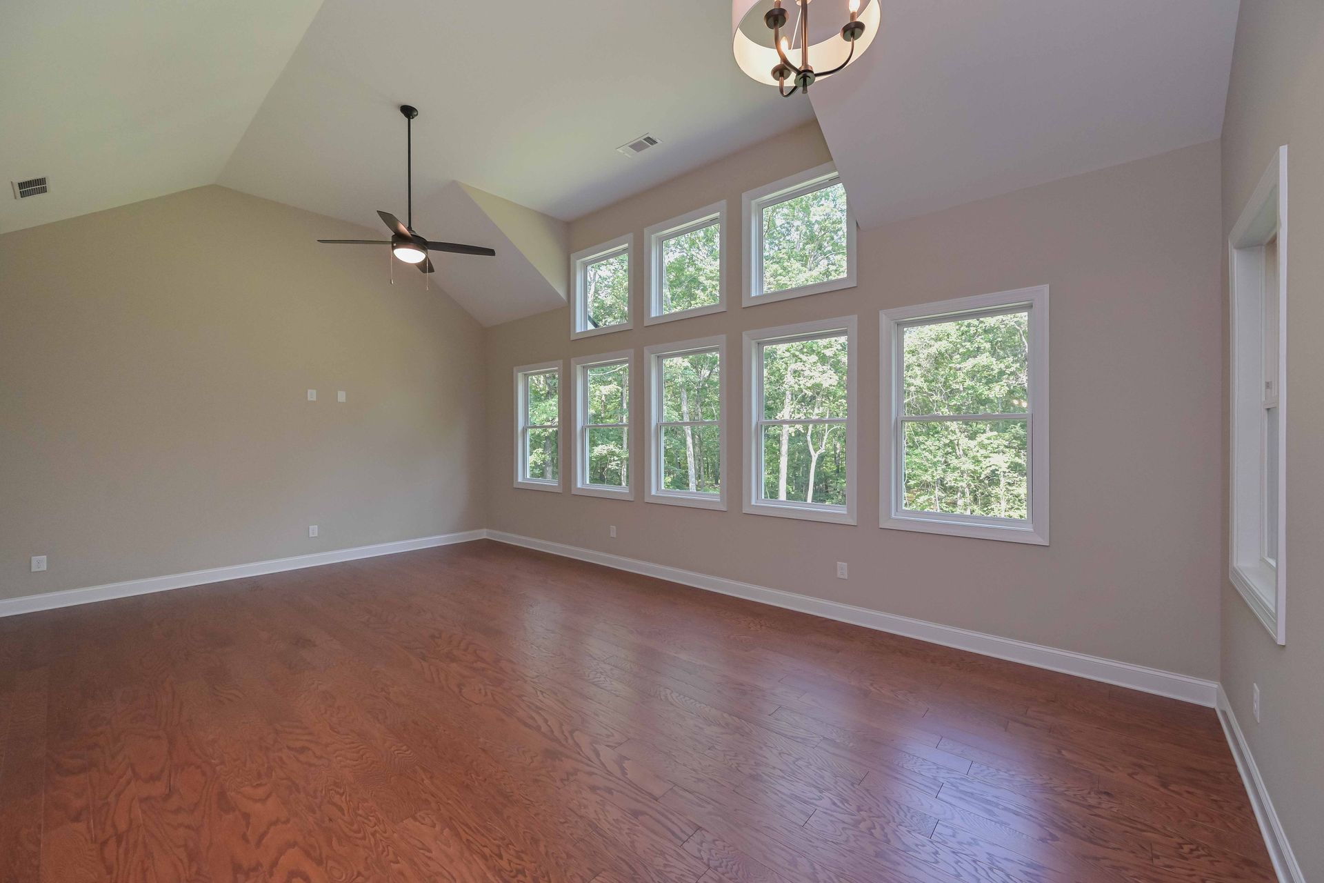 Empty living room with hardwood floors, high ceiling, ceiling fan, and large windows overlooking trees