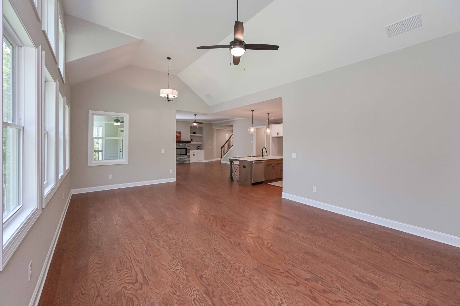 Empty open-plan living room with wood floors, vaulted ceiling, ceiling fan, and kitchen visible in back