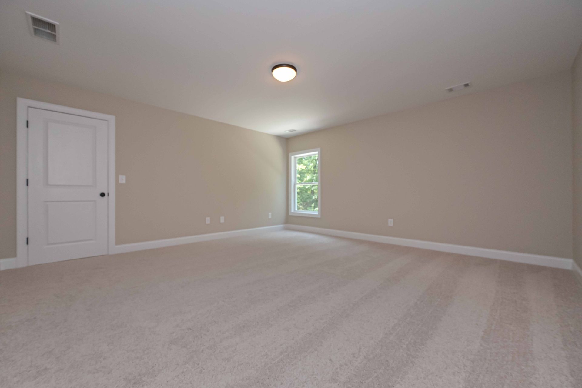 Empty beige carpeted bedroom with white door, small window, and ceiling light