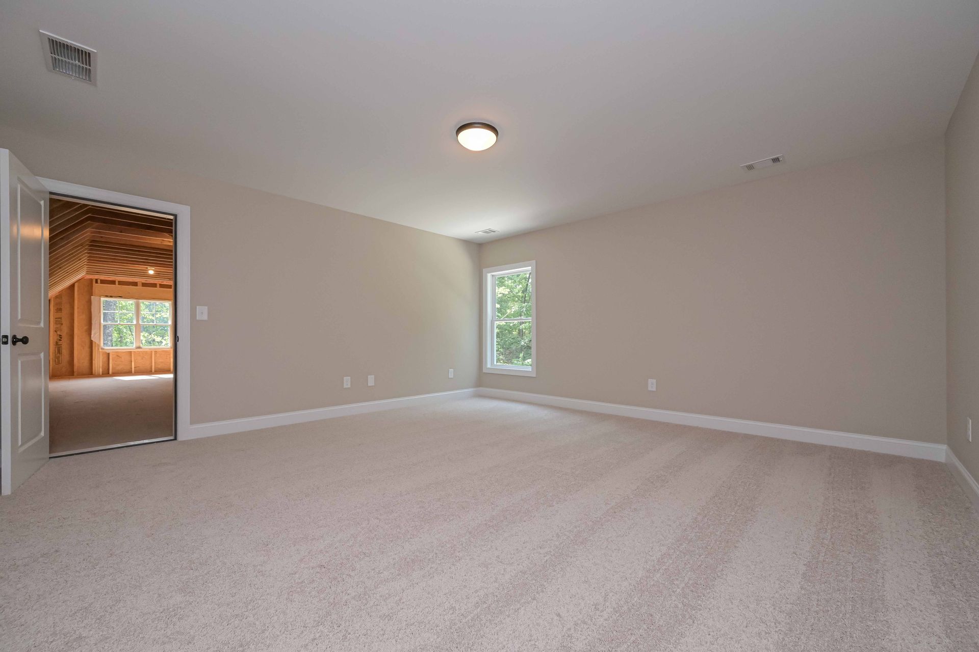 Empty beige carpeted room with white walls, a ceiling light, and a doorway to a sunlit space