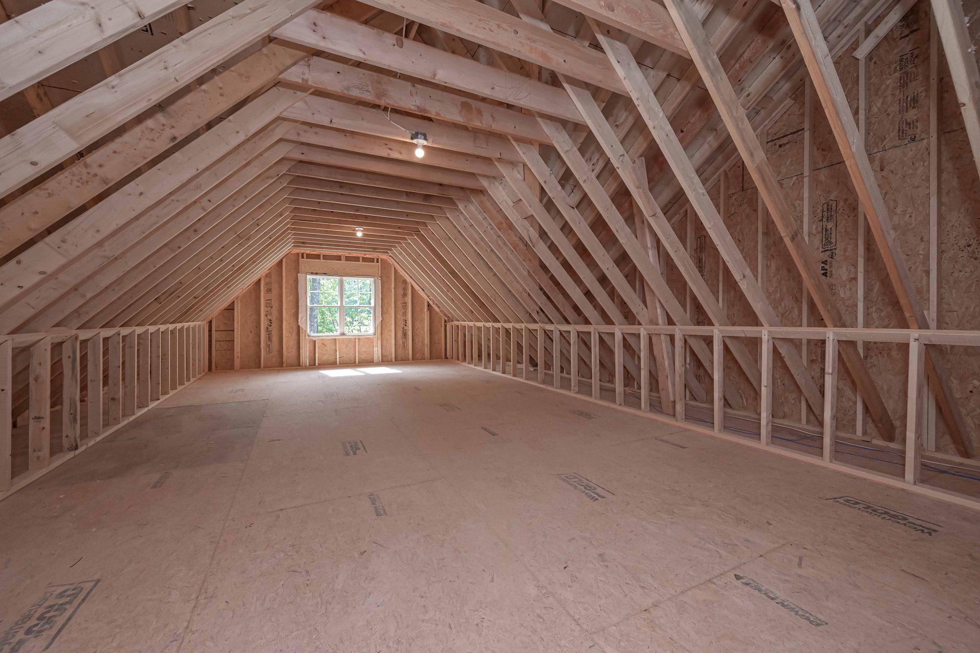 Unfinished attic interior with exposed wooden trusses, insulation, and a small window at the far end.