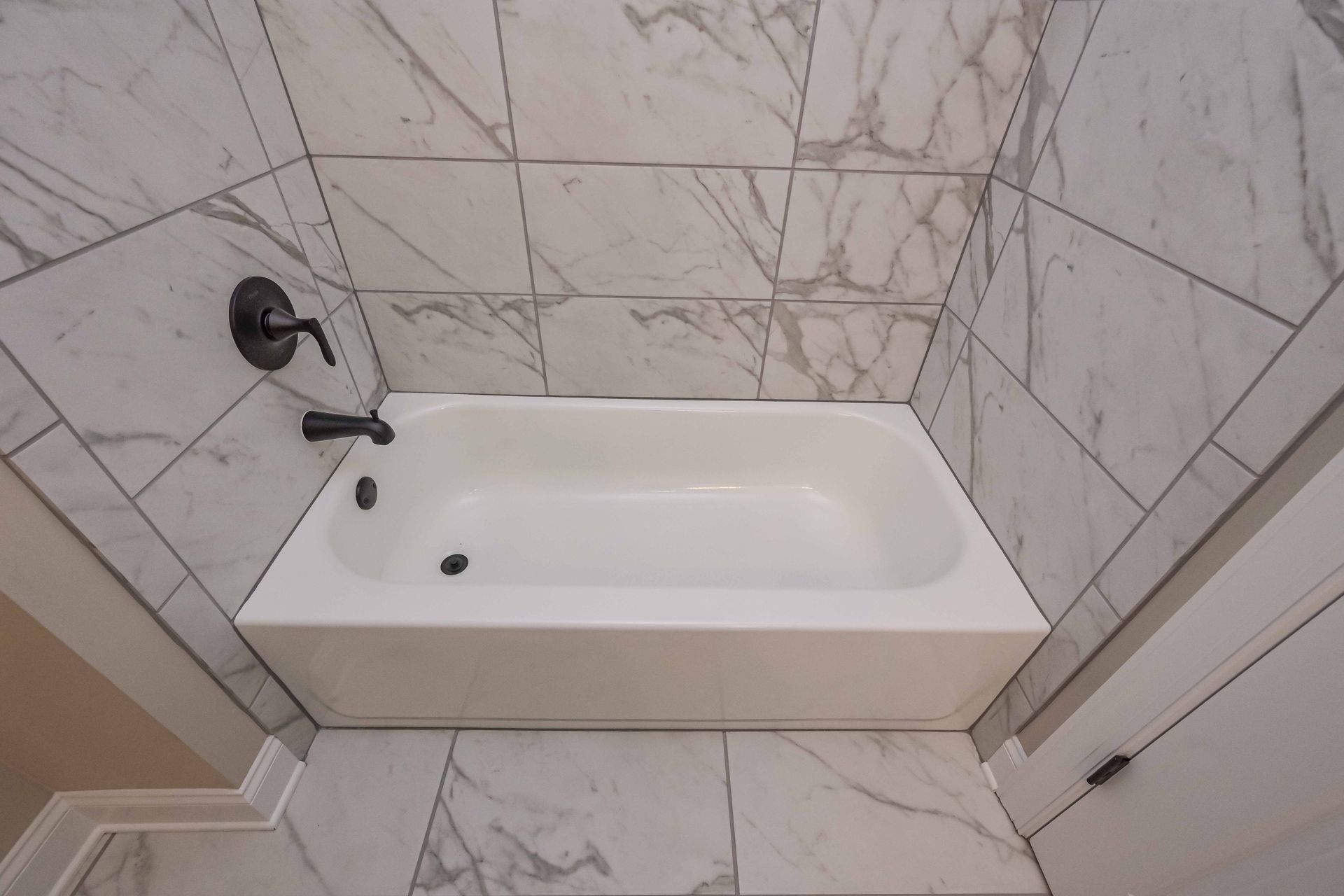 White bathtub in a marble-tiled bathroom with a black wall-mounted faucet and showerhead.