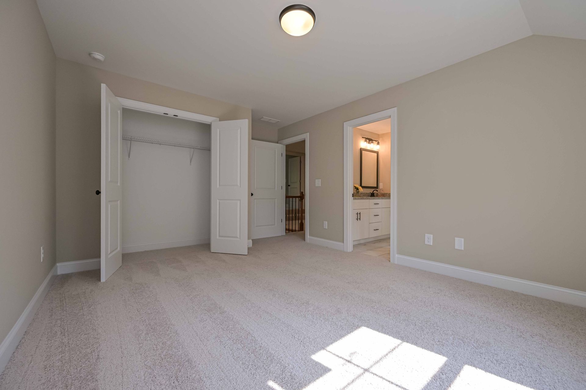 Empty beige bedroom with open closet and bathroom door, carpeted floor, and sunlight on the floor
