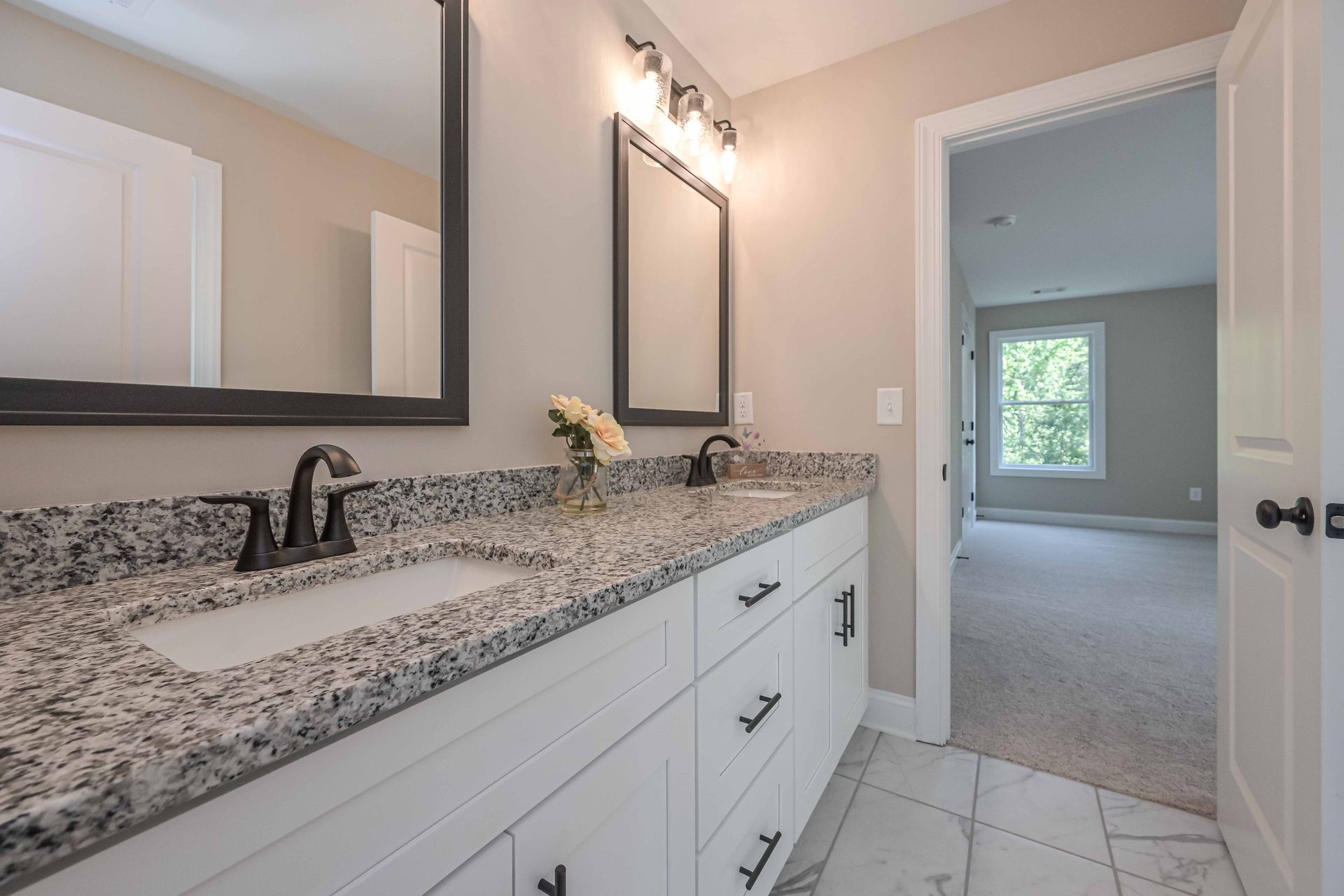 Bright bathroom vanity with white cabinets, granite countertop, and large mirror leading to a carpeted room.