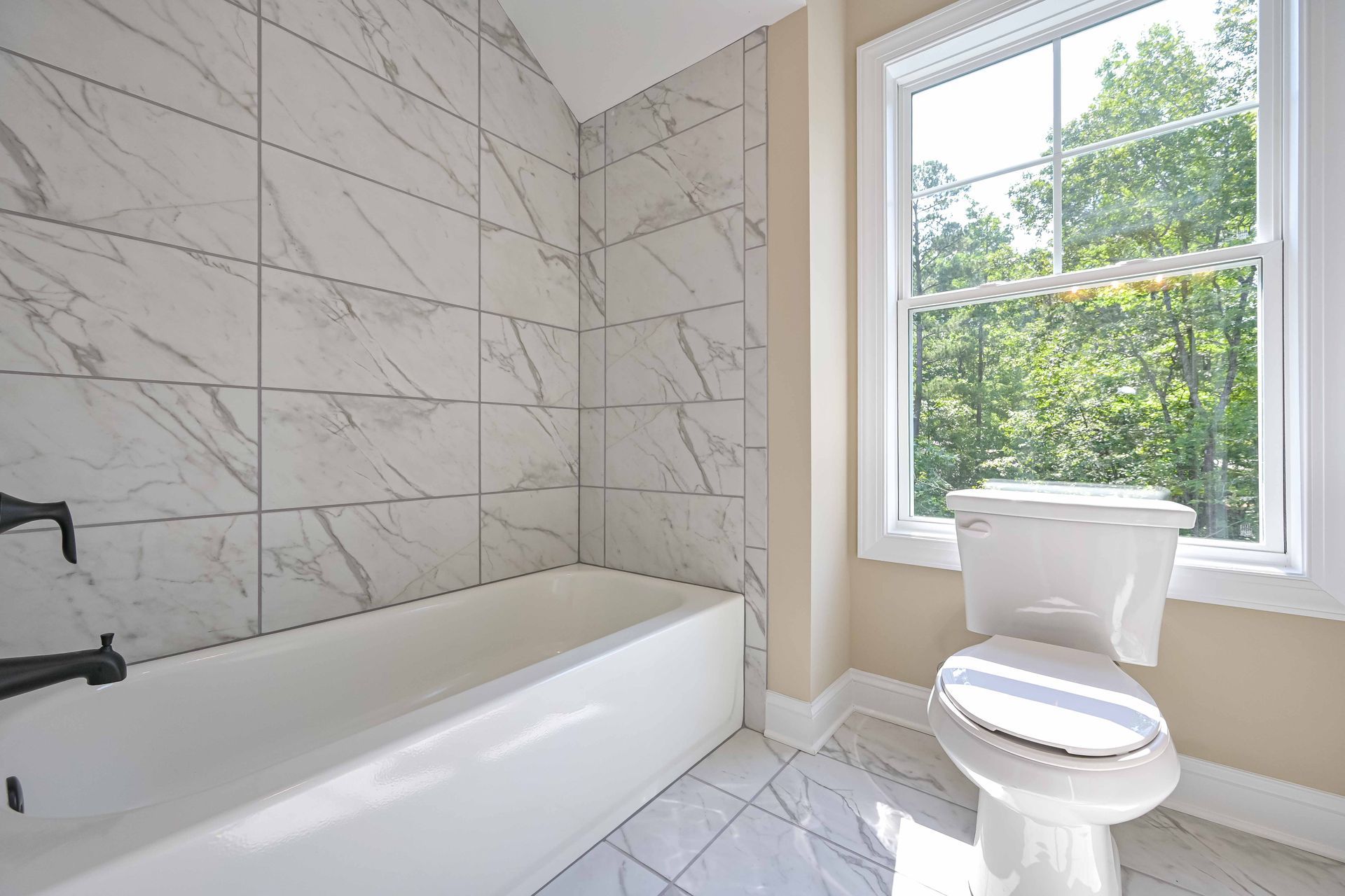 Bright bathroom with marble-tiled tub, white toilet, and a window overlooking greenery