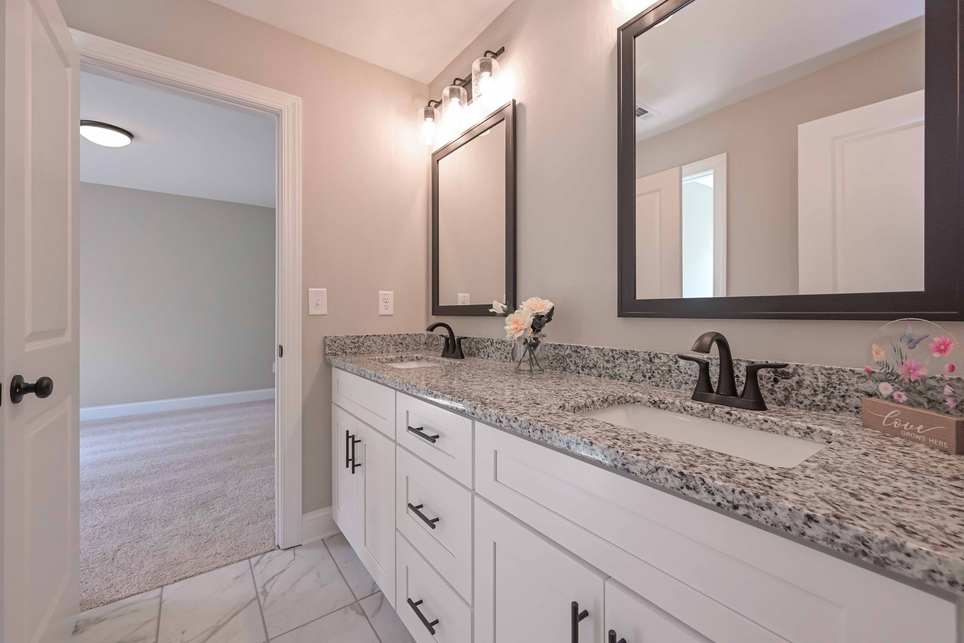 Bright bathroom vanity with granite countertop, double sinks, mirrors, and tiled floor; open doorway to carpeted room.