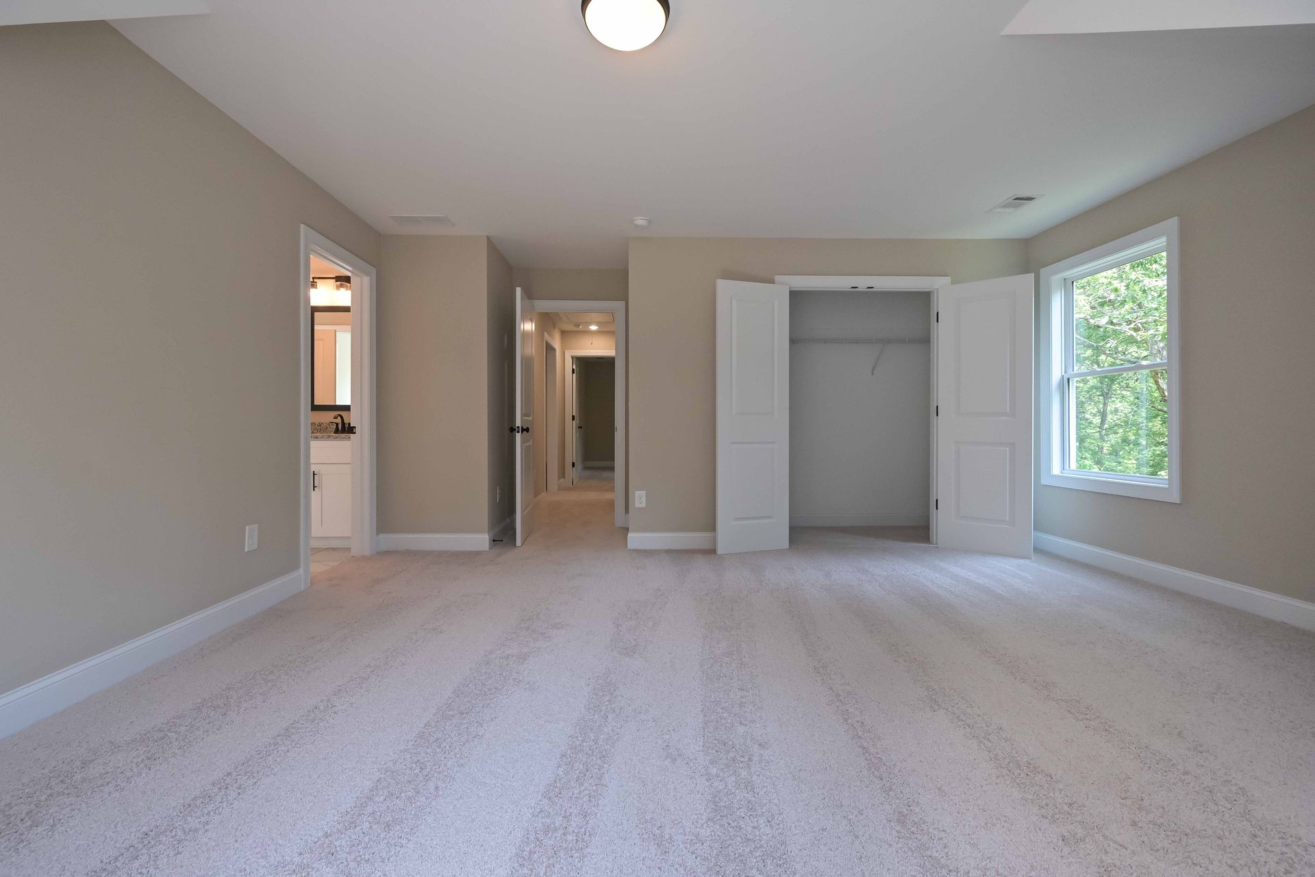 Empty beige bedroom with carpet, closet doors, and a window showing greenery.
