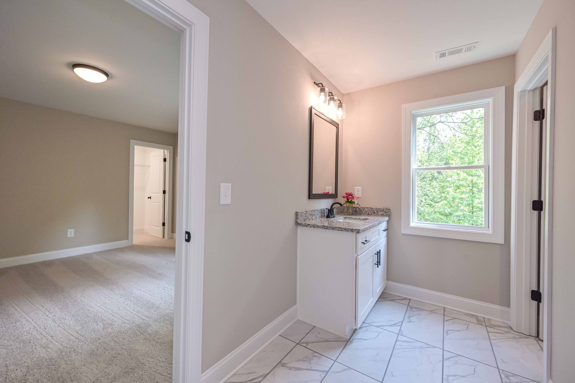 Bright bathroom with white vanity, tiled floor, mirror, and window, opening to a carpeted room.