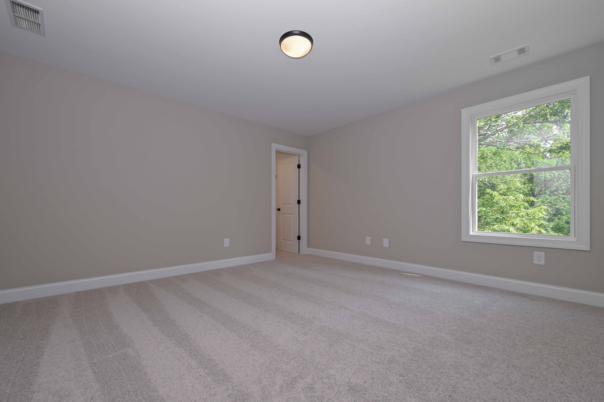 Empty beige bedroom with gray carpet, white trim, a ceiling light, and a window with green trees outside