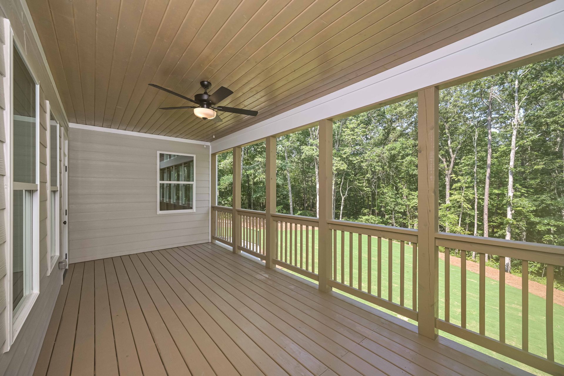 Covered wooden porch with railing and ceiling fan overlooking a green wooded yard.