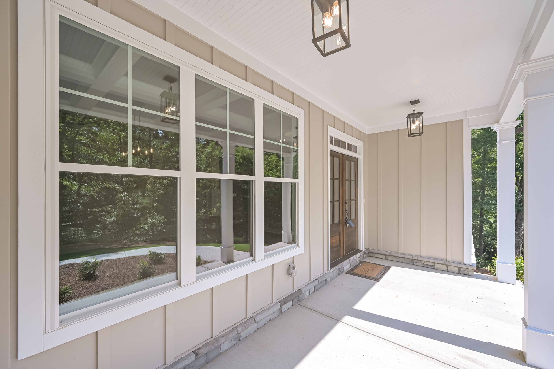 Covered front porch with white trim, large windows, tan siding, and a bright concrete floor