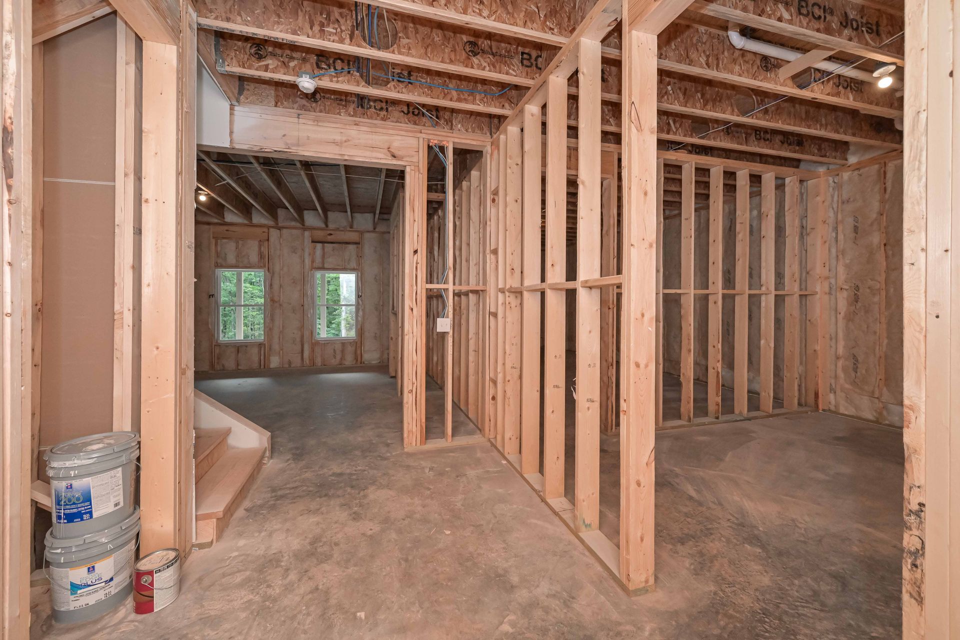 Unfinished basement framing with exposed studs, concrete floor, and windows at the far end
