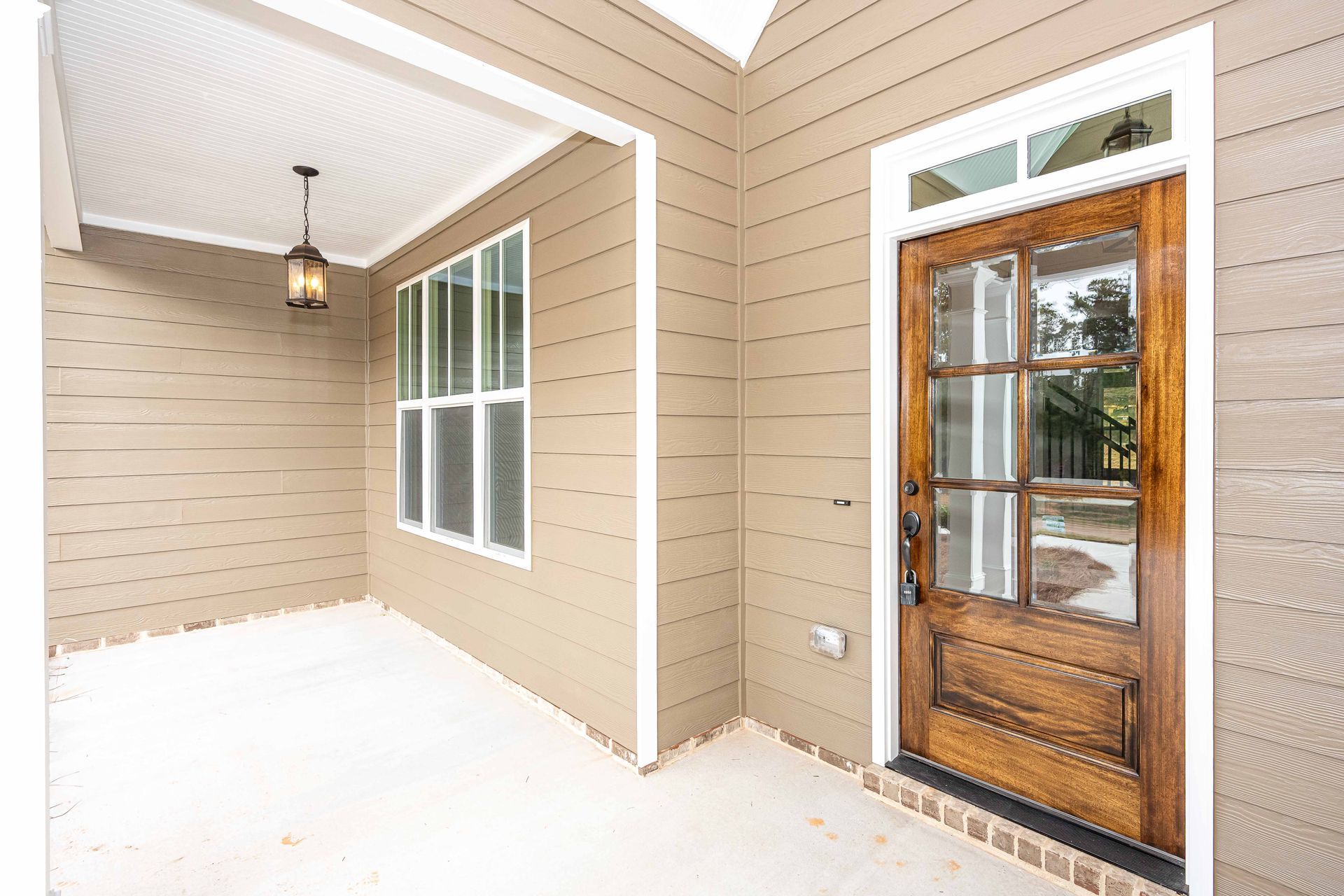 Covered front porch with beige siding, white ceiling, hanging light, window, and a wooden glass-paneled door.
