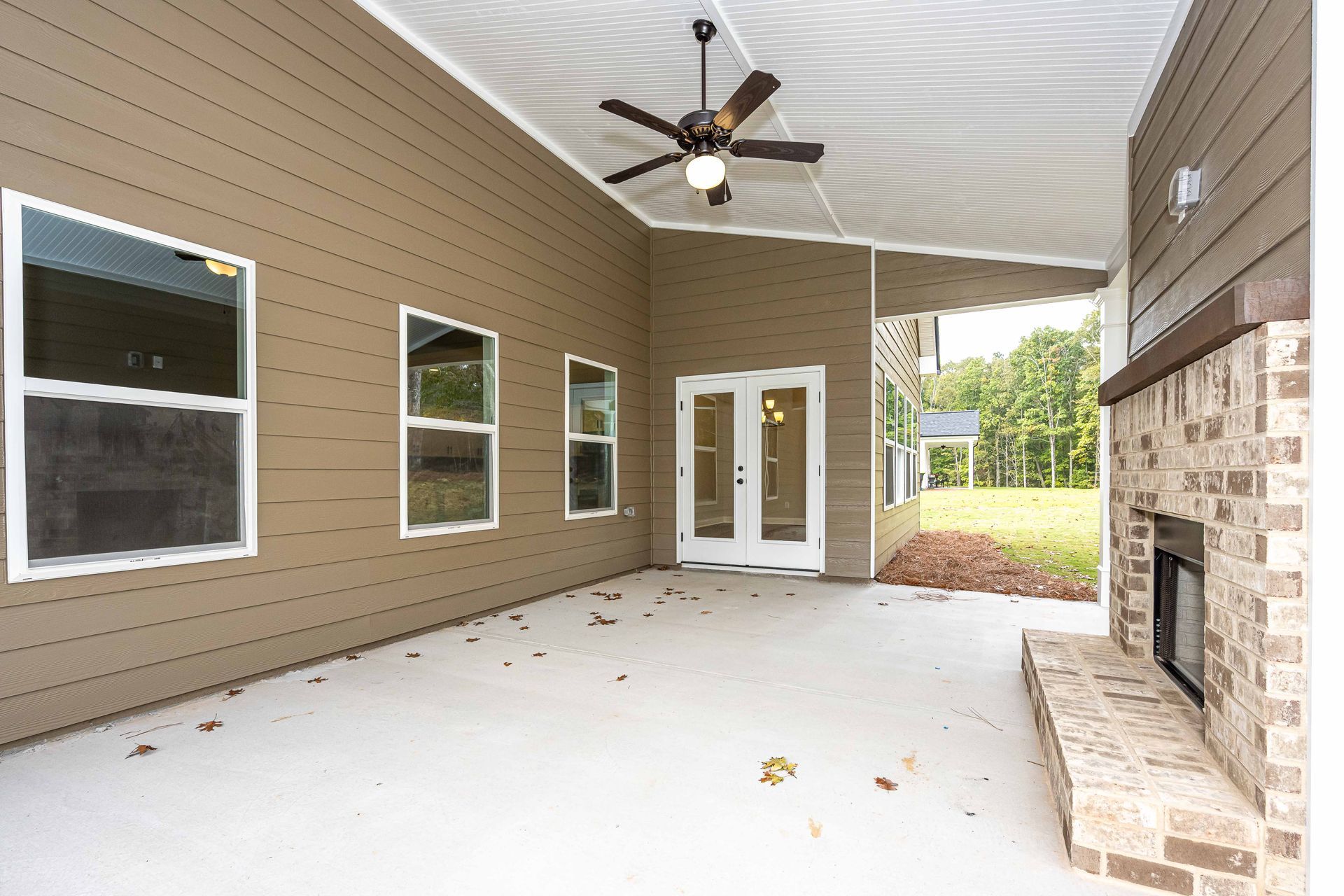 Covered patio with ceiling fan, white concrete floor, tan siding, windows, glass doors, and brick fireplace
