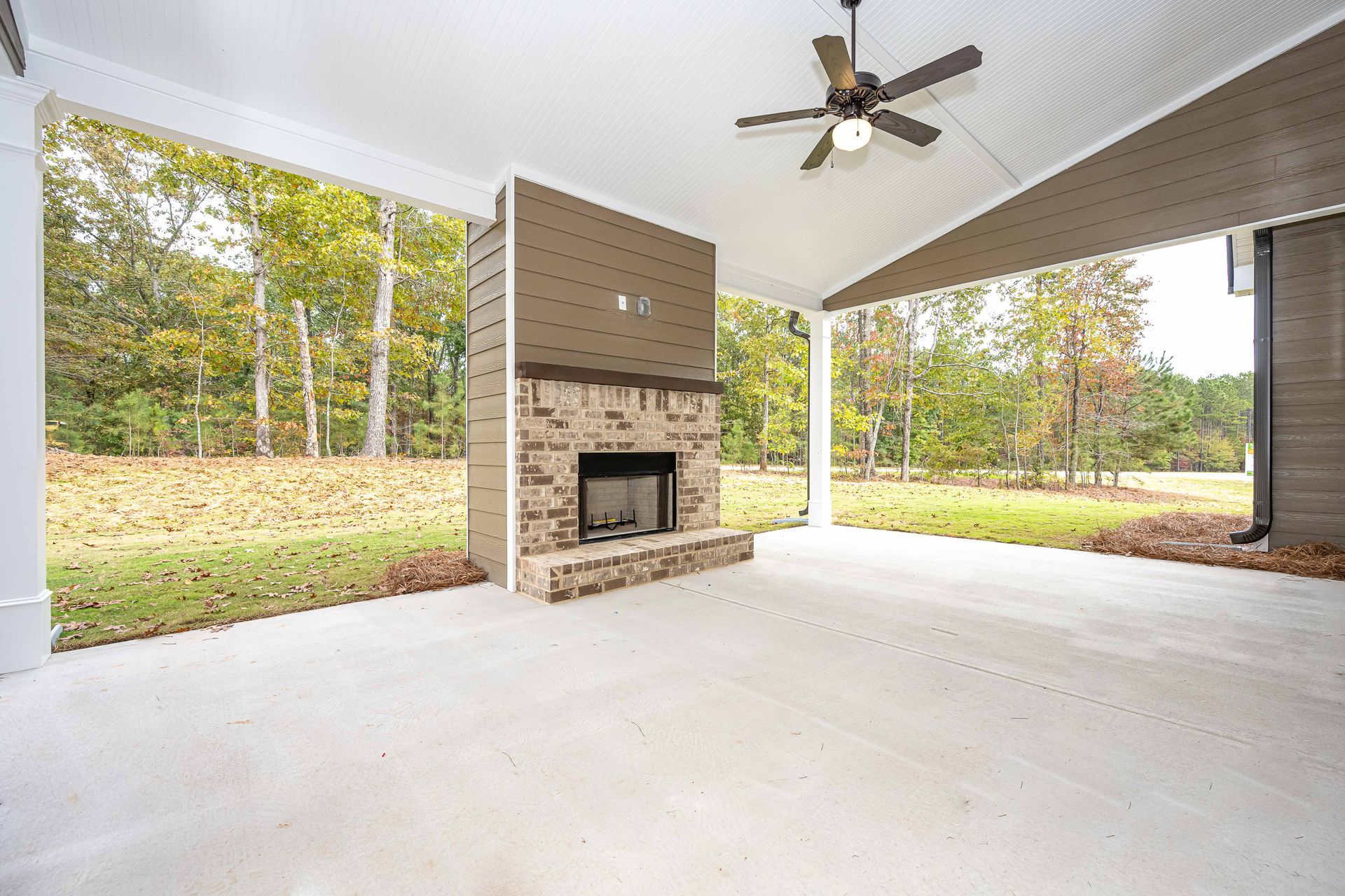 Covered patio with a brick fireplace, ceiling fan, and open view of a grassy yard and trees