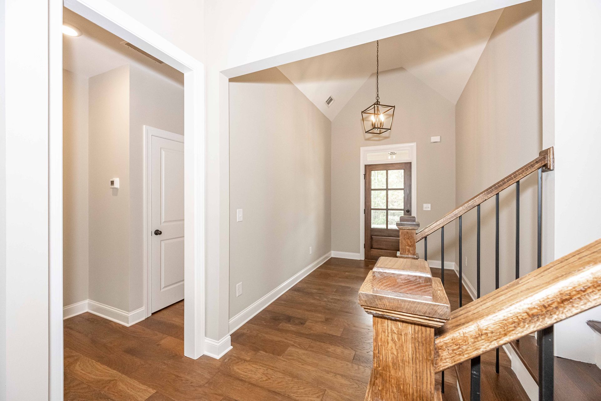 Bright hallway with hardwood floors, staircase railing, and hanging light leading to a windowed room.