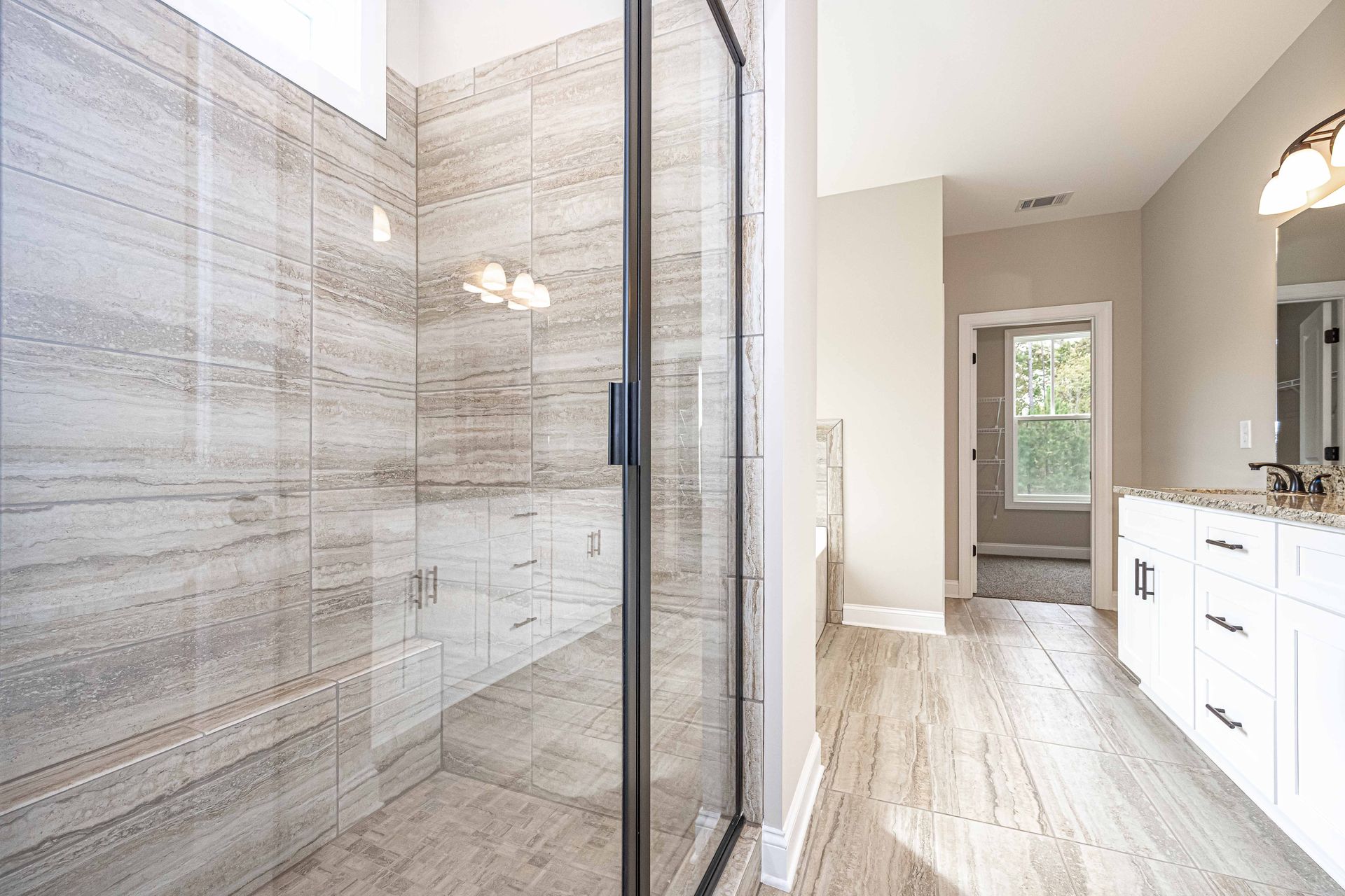 Modern bathroom with glass shower, marble tile, and white vanity leading to a bedroom doorway