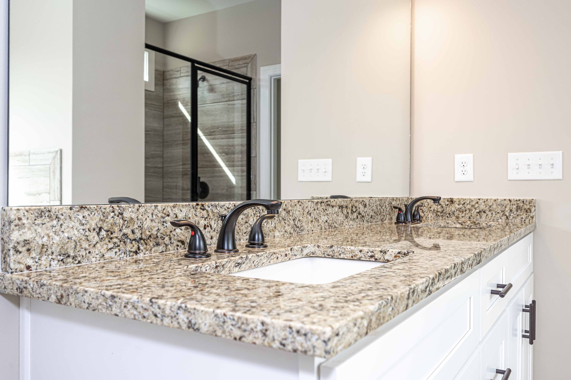 Modern bathroom vanity with granite countertop, dual faucets, and a white sink near a glass shower door