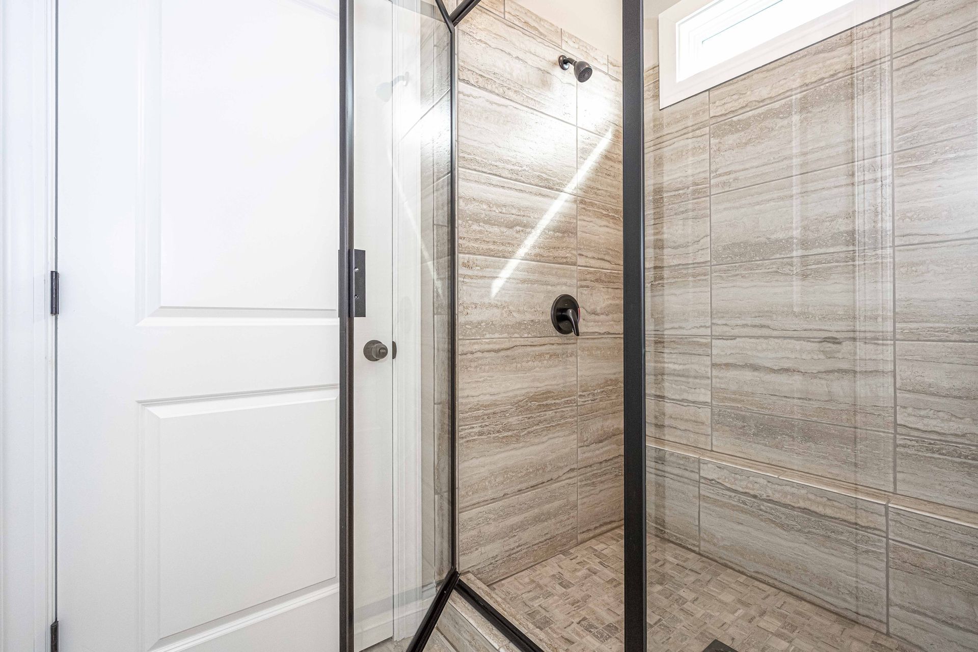 Modern tiled shower with glass enclosure and adjacent white door, viewed in a bright bathroom
