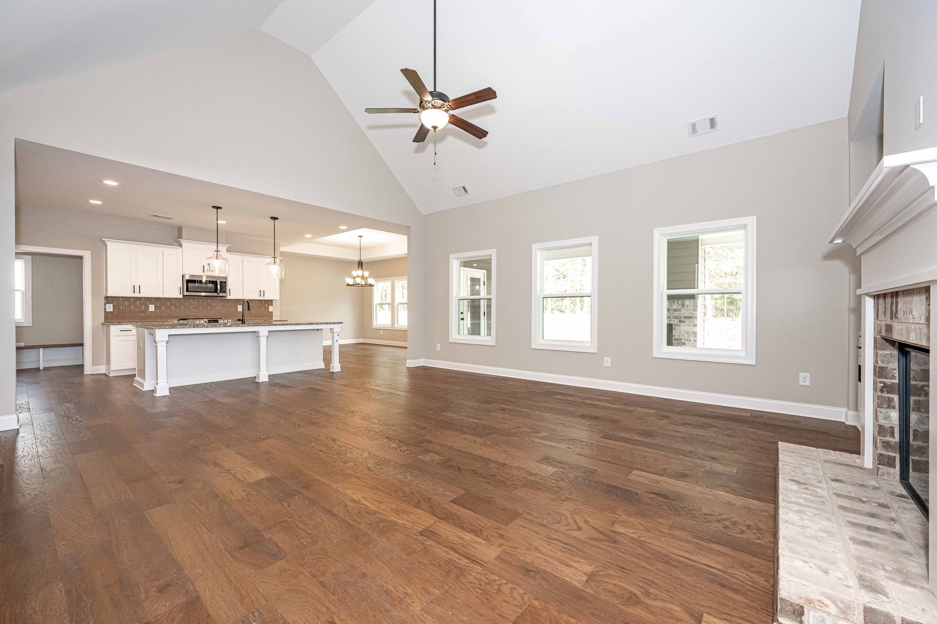 Open-concept living room with hardwood floors, vaulted ceiling, ceiling fan, and fireplace beside a bright kitchen