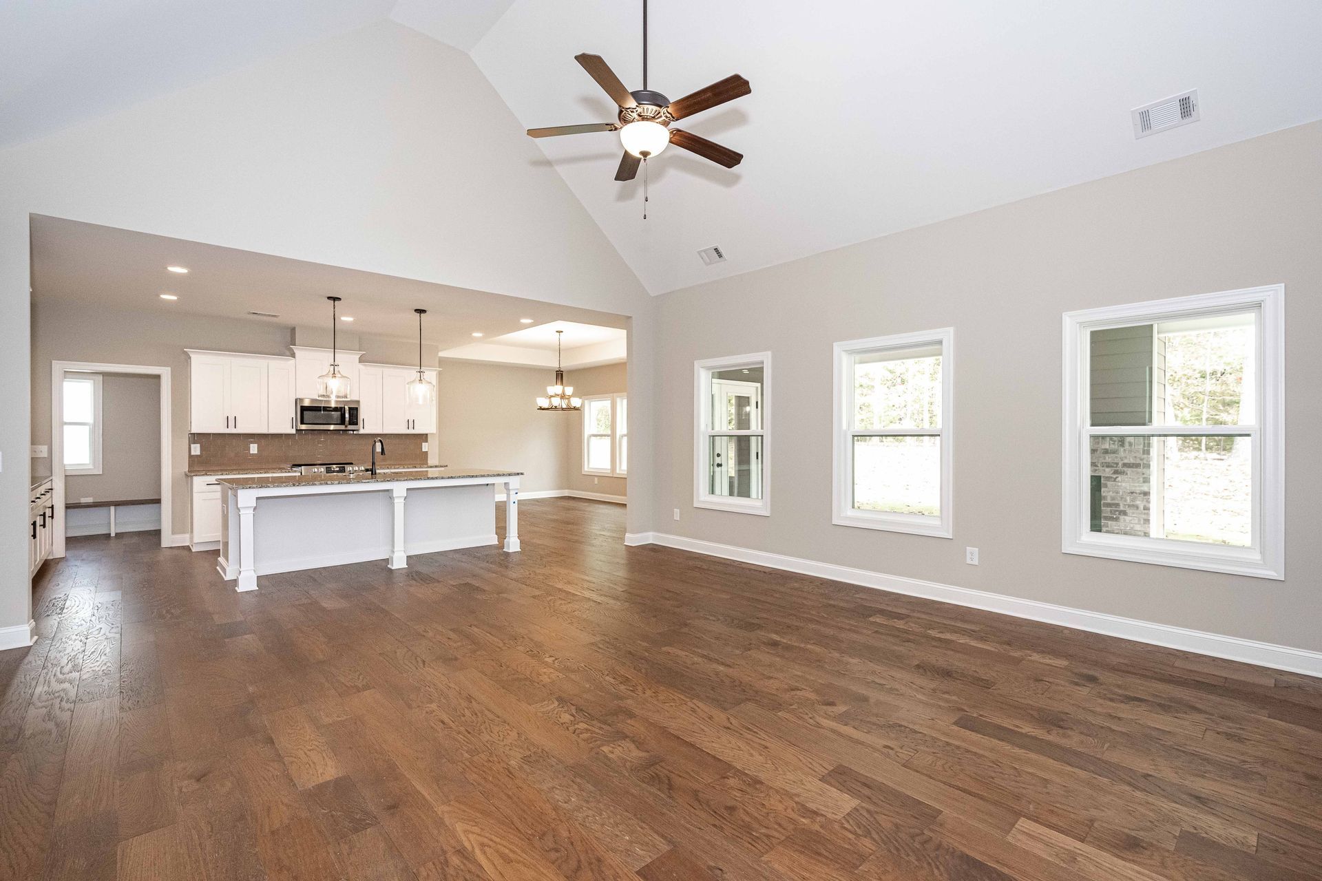 Spacious empty living room with hardwood floors, white walls, large windows, and an open kitchen with island.
