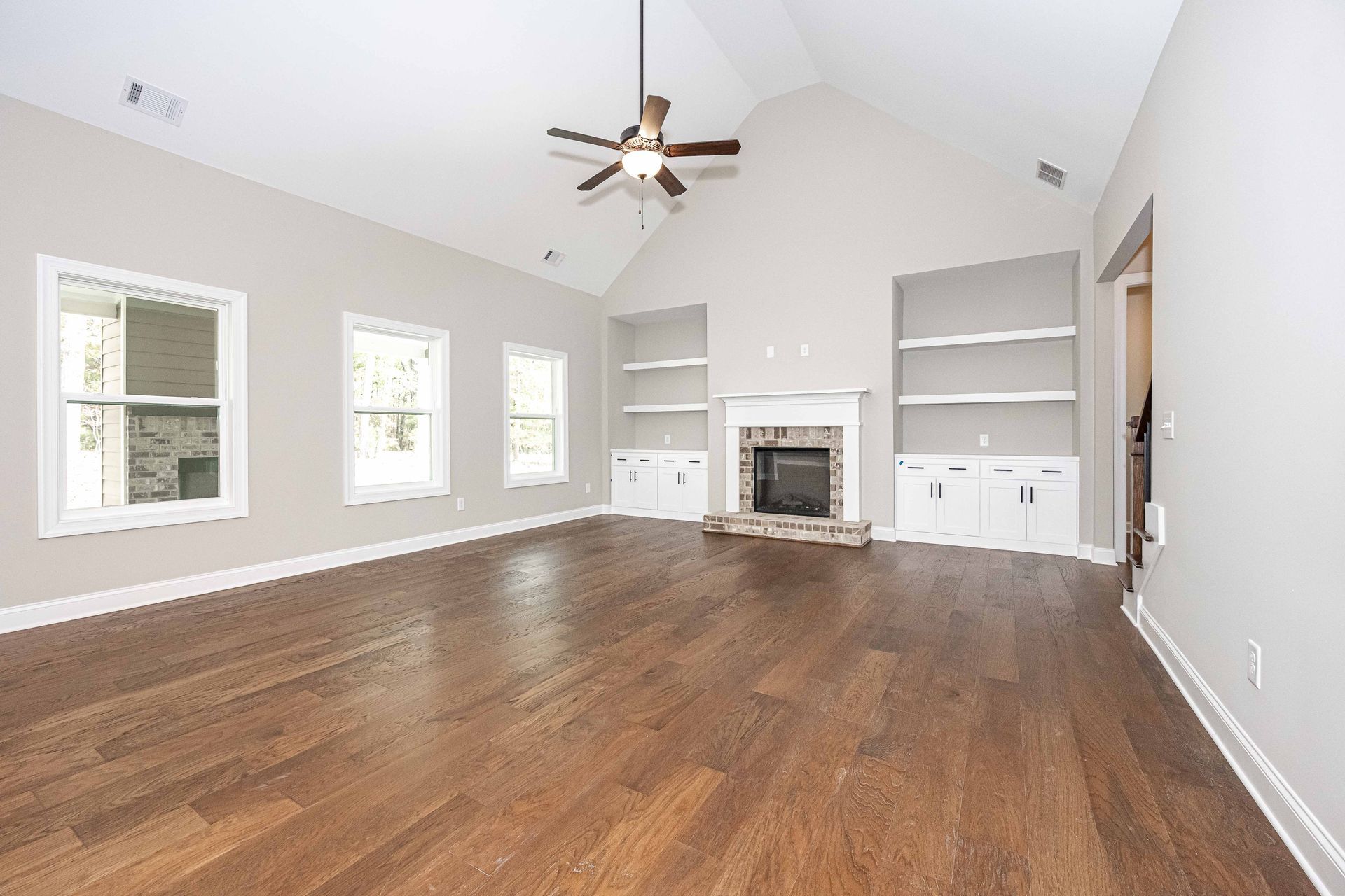 Spacious empty living room with hardwood floors, vaulted ceiling, ceiling fan, fireplace, and white built-ins.
