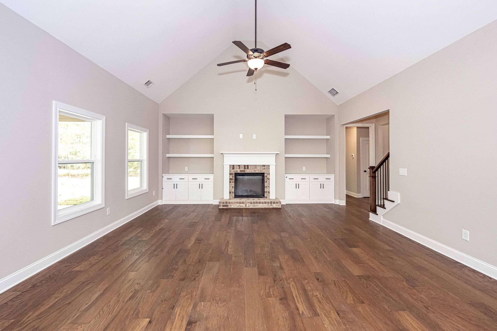 Spacious empty living room with vaulted ceiling, ceiling fan, fireplace, built-in shelves, and wood floors