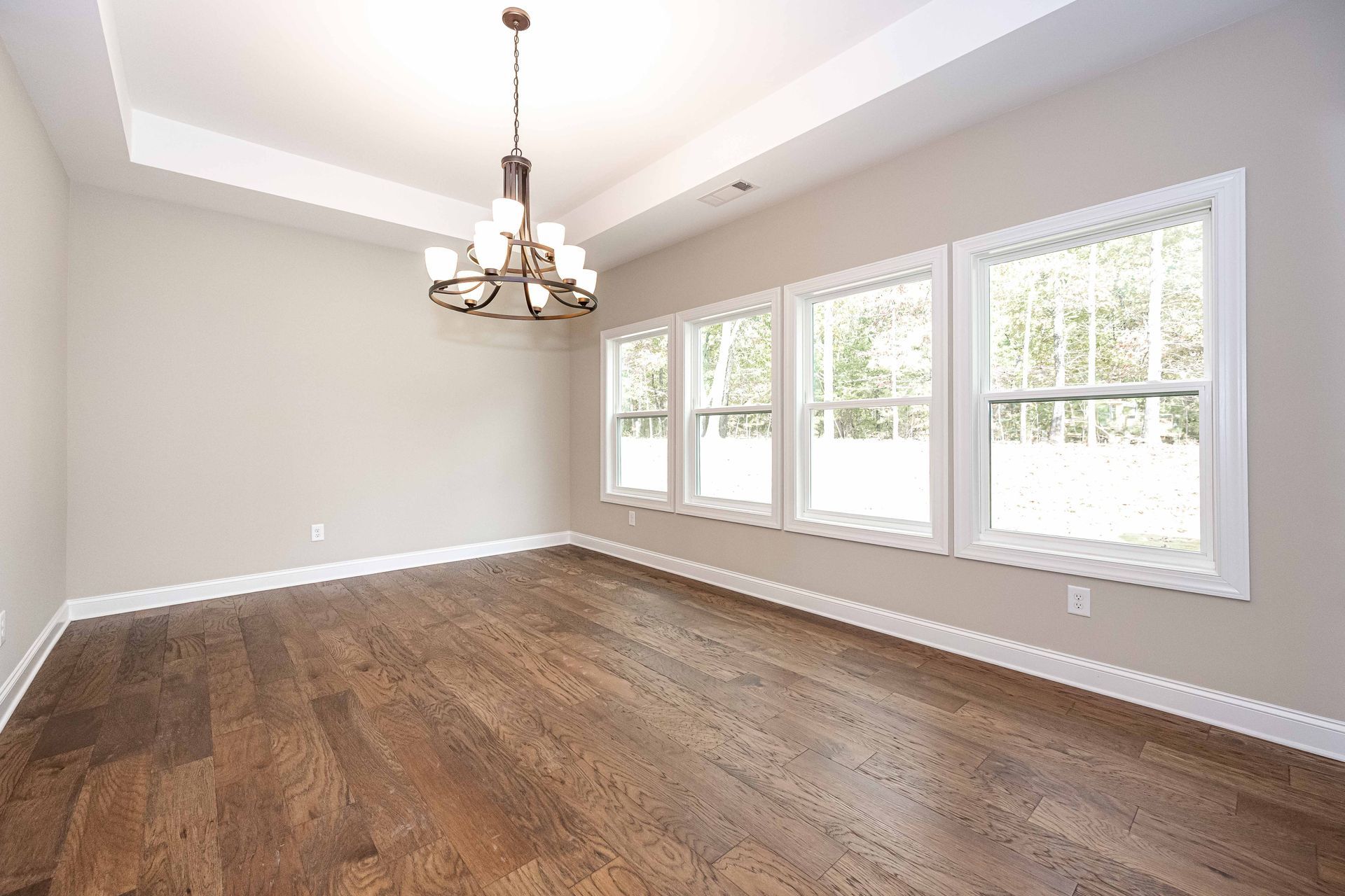 Empty living room with hardwood floors, beige walls, chandelier, and large sunlit windows.