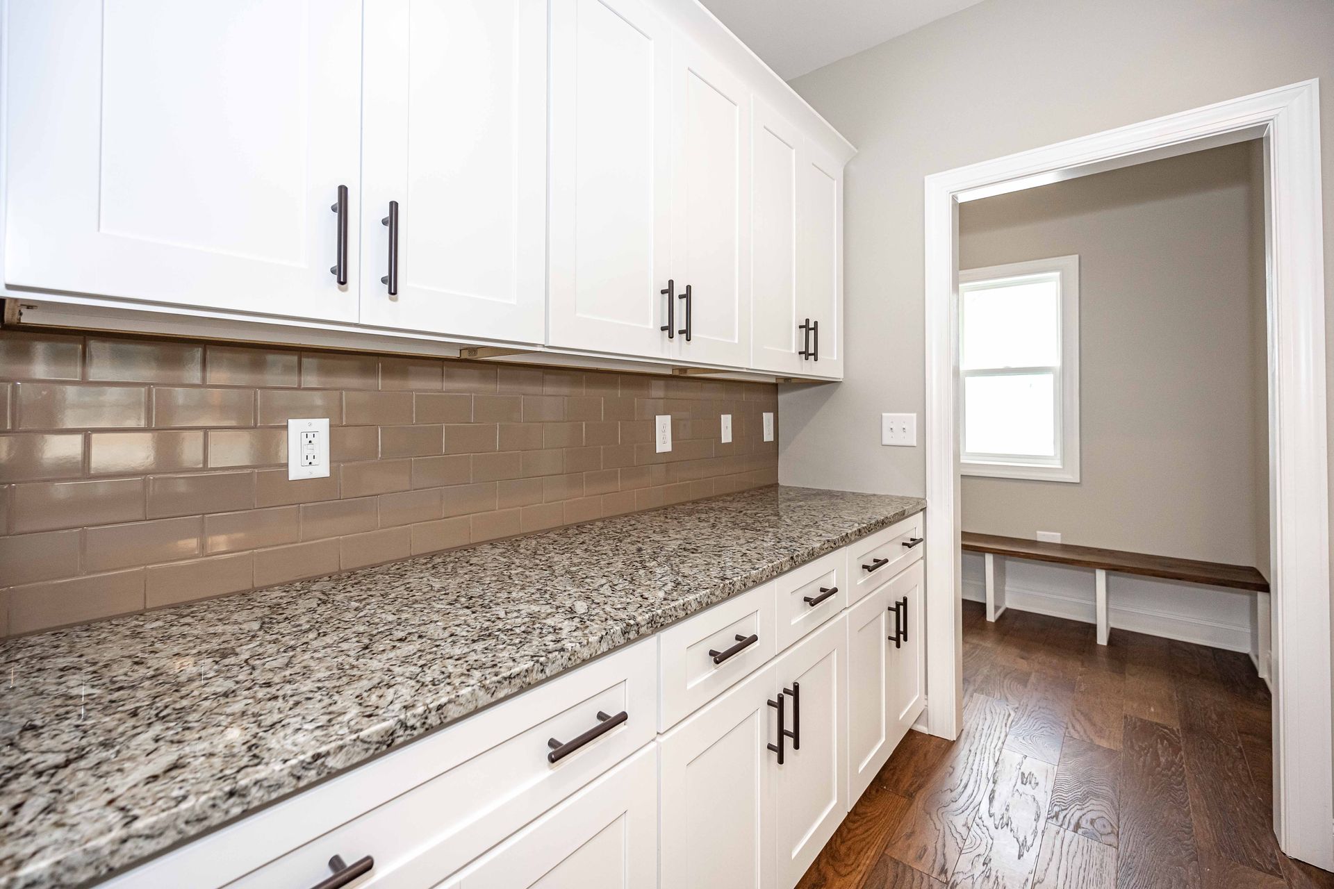 Bright kitchen with white cabinets, speckled countertop, and brown tile backsplash leading into a dining area.