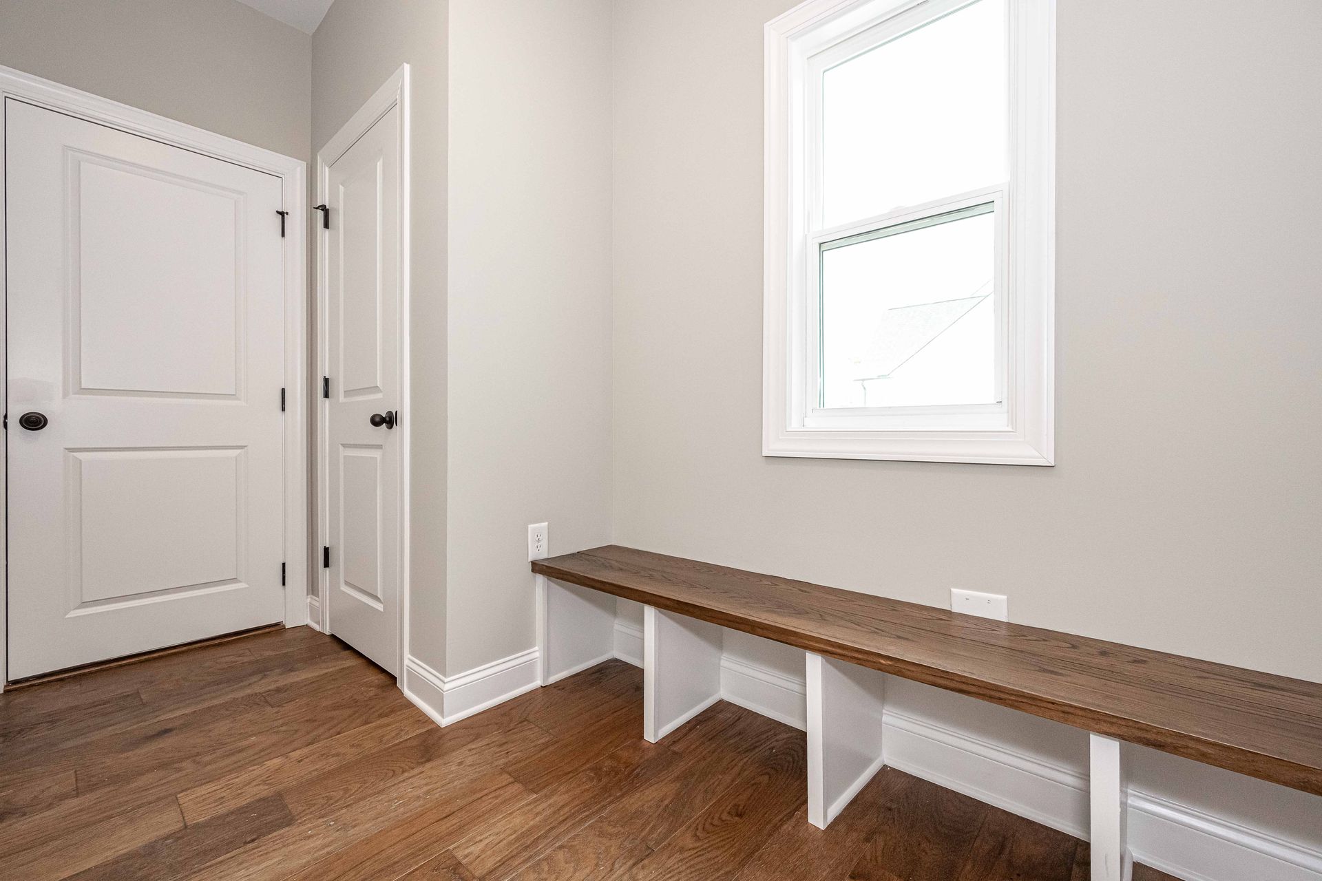Empty hallway with white doors, wood floor, and a long bench beneath a window