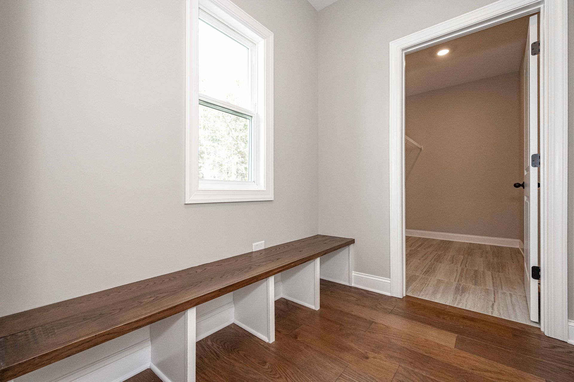 Minimal hallway with a long wooden bench, small window, and open door to a carpeted room.