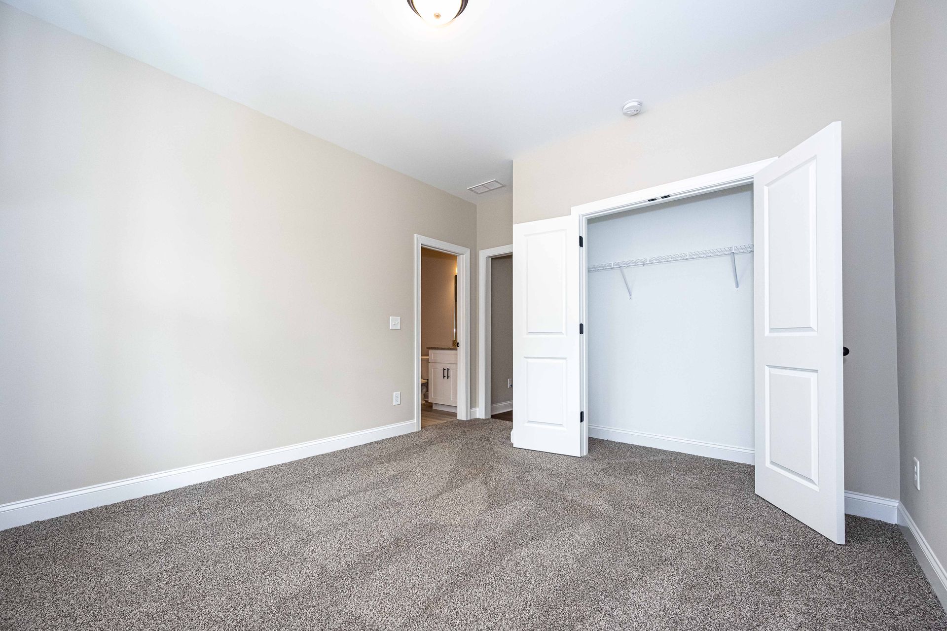 Empty bedroom with beige walls, carpeted floor, open closet doors, and a doorway to the hall.