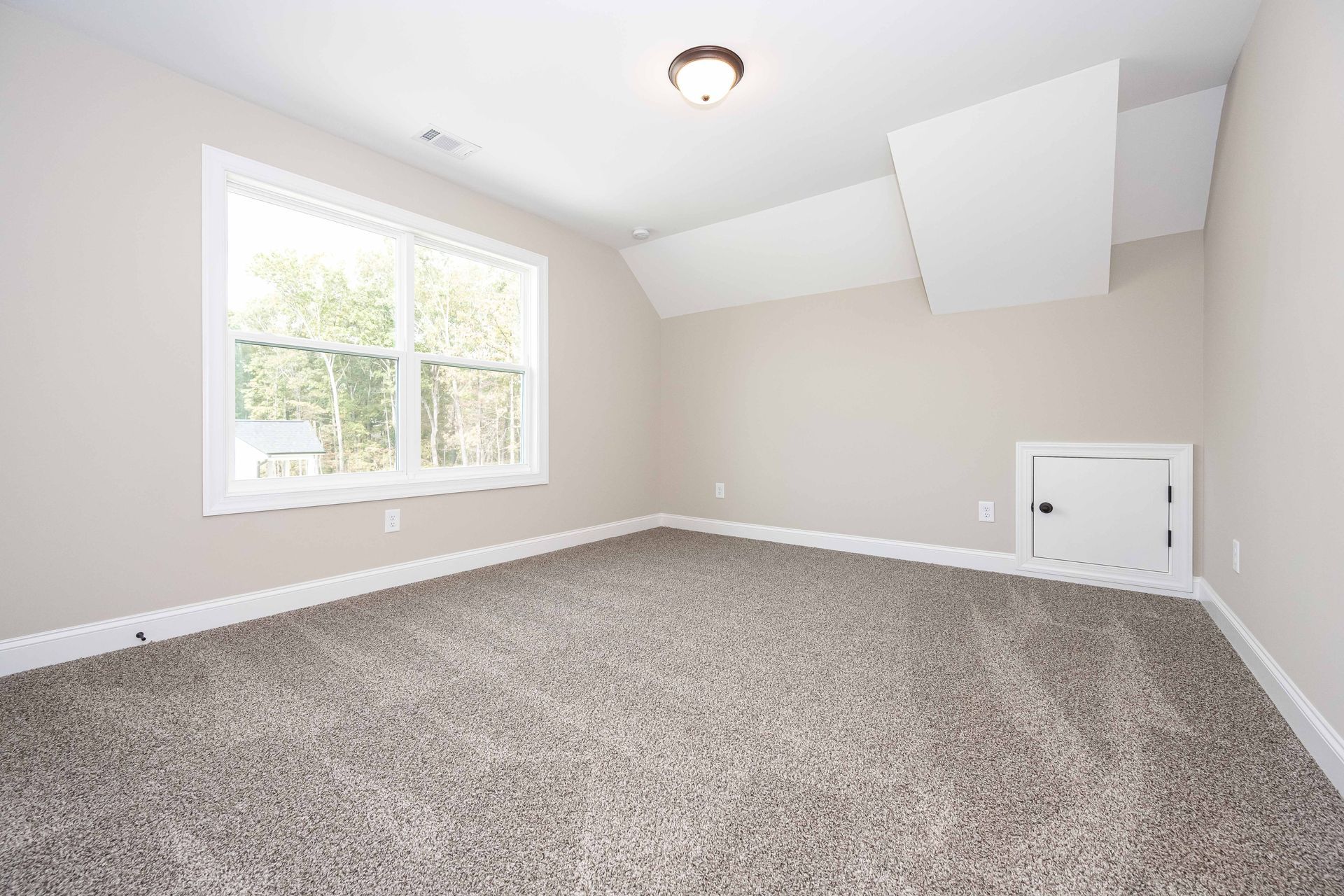 Empty beige bedroom with carpet, window, and a small attic access door on the right