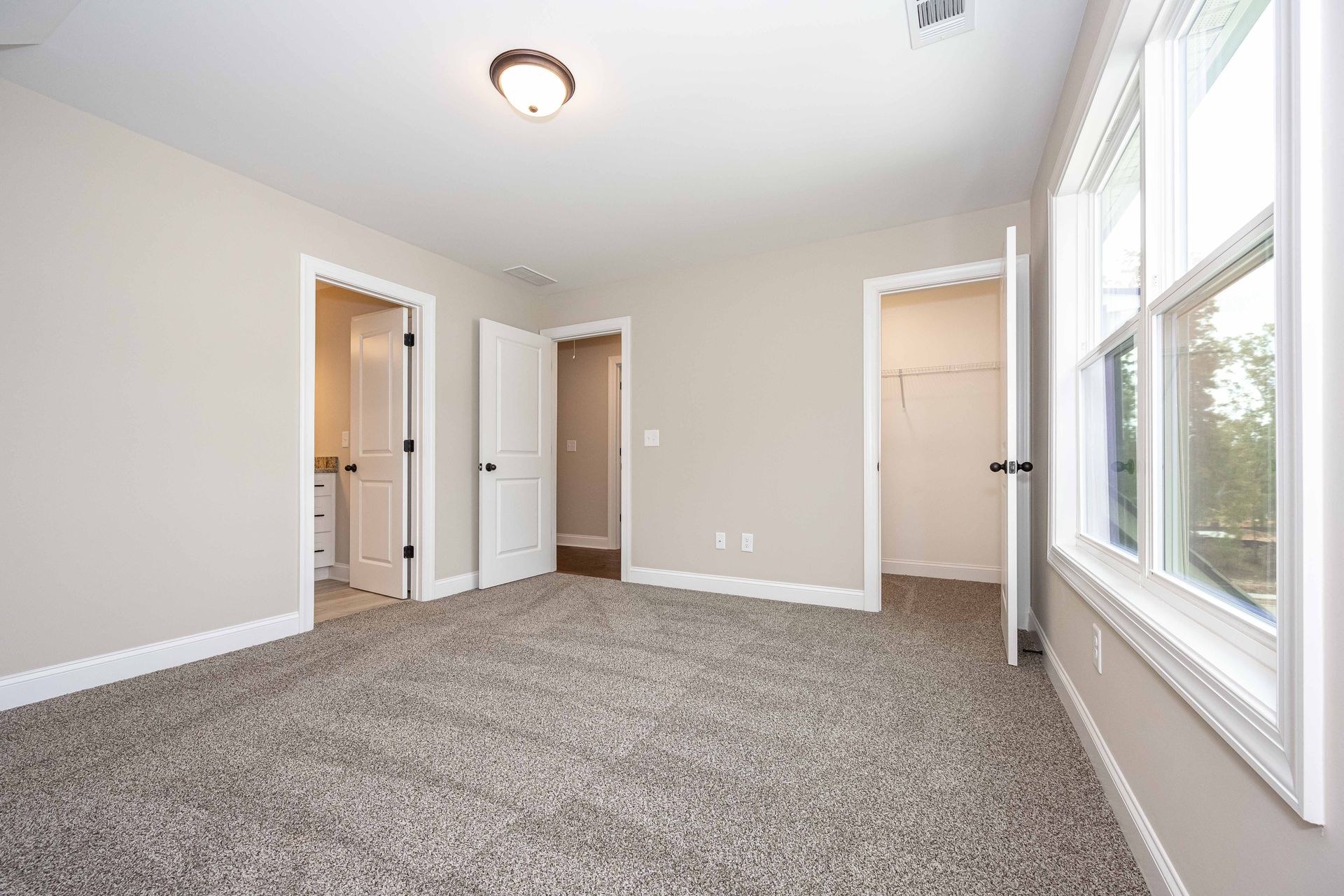 Empty beige bedroom with carpet, white trim, and large windows letting in daylight.