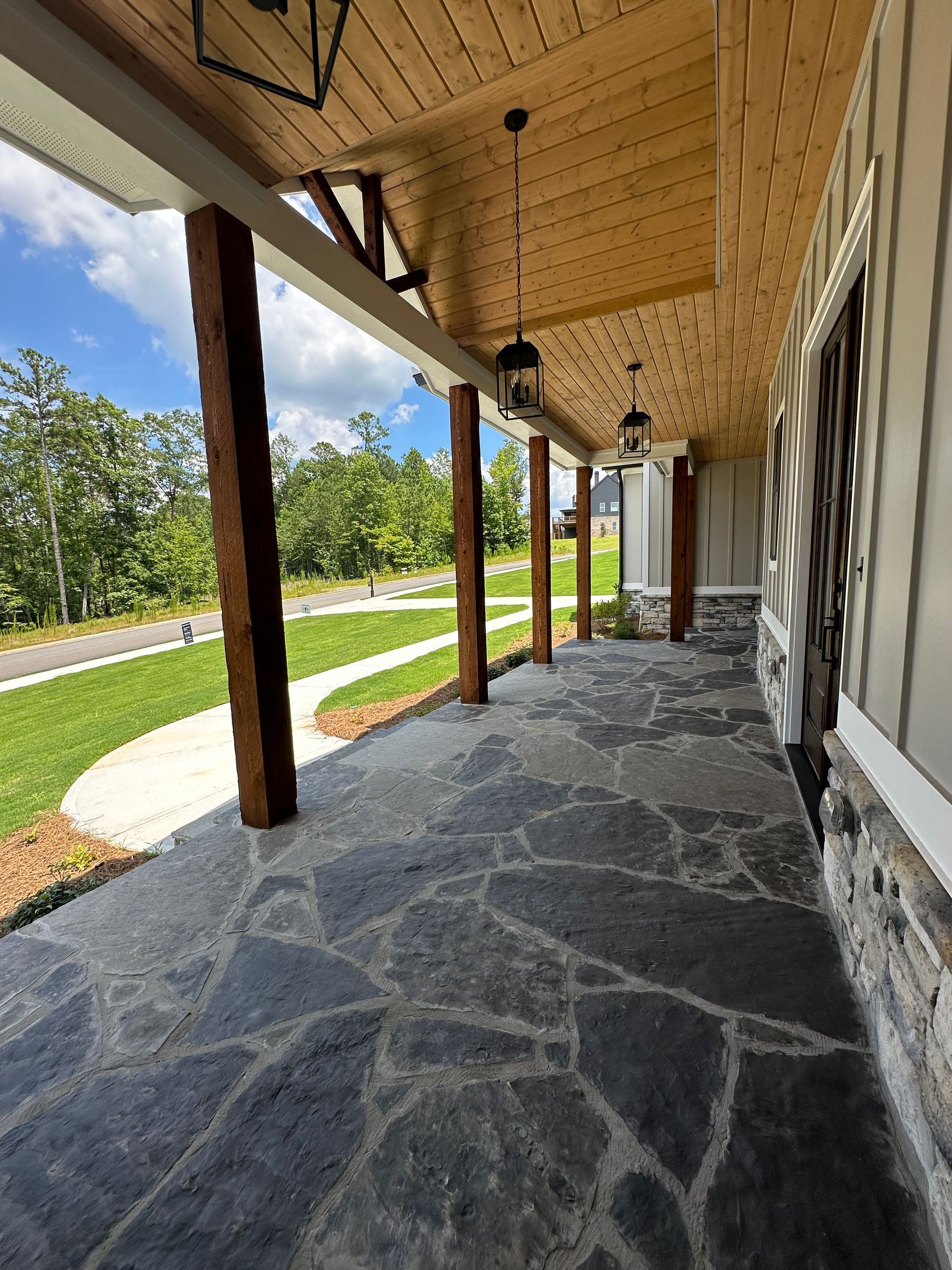 Covered stone porch with wooden posts, hanging lanterns, and a view of a grassy lawn and trees