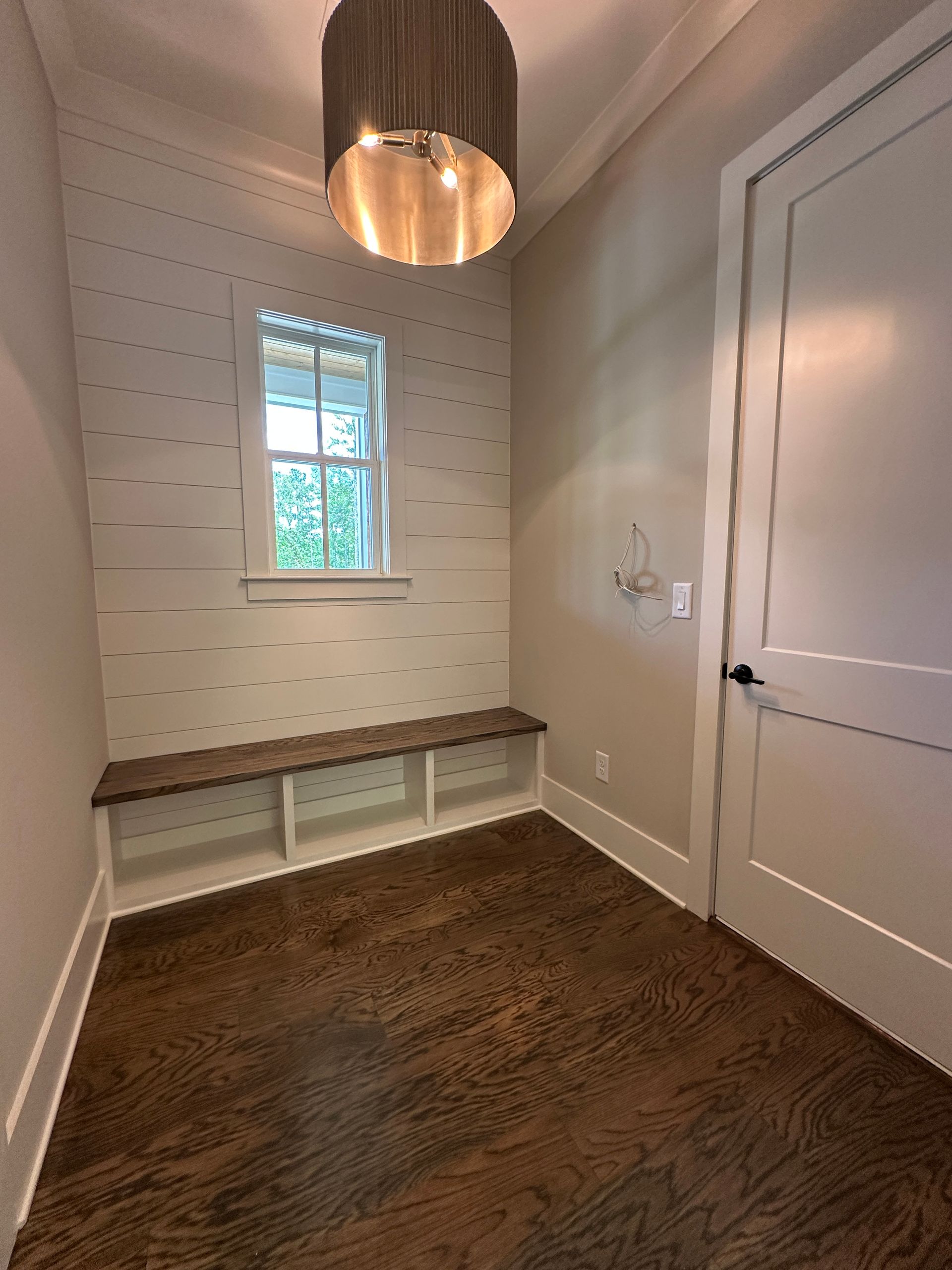 Small hallway nook with white walls, bench seating, a window, and a hanging pendant light over wood flooring.