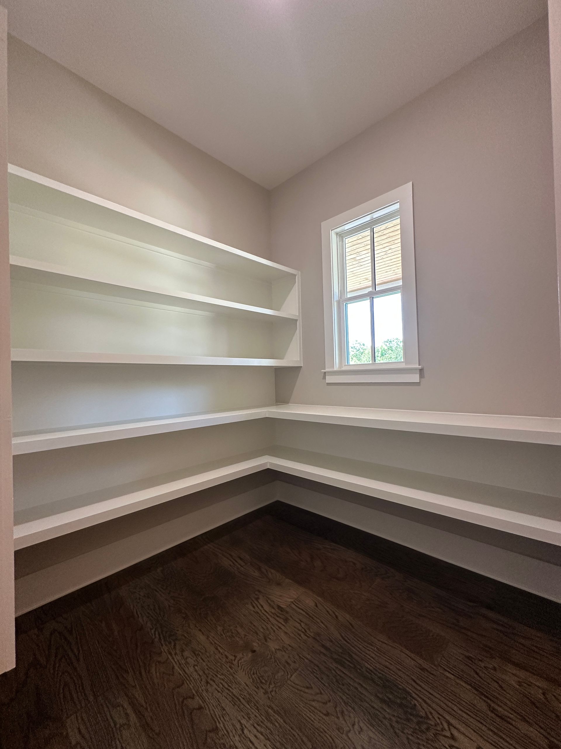 Empty walk-in closet with white built-in shelves, dark wood floor, and a small window.