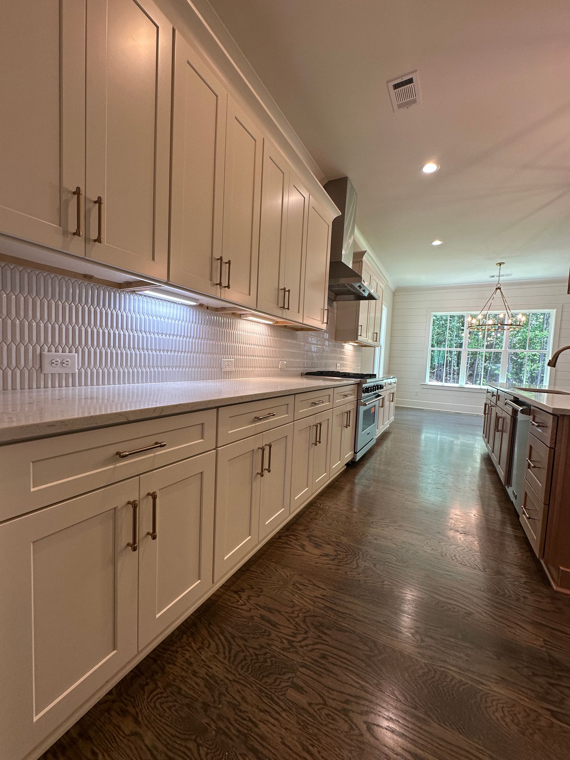Bright white galley kitchen with long cabinets, tiled backsplash, and a view of greenery through the far window.