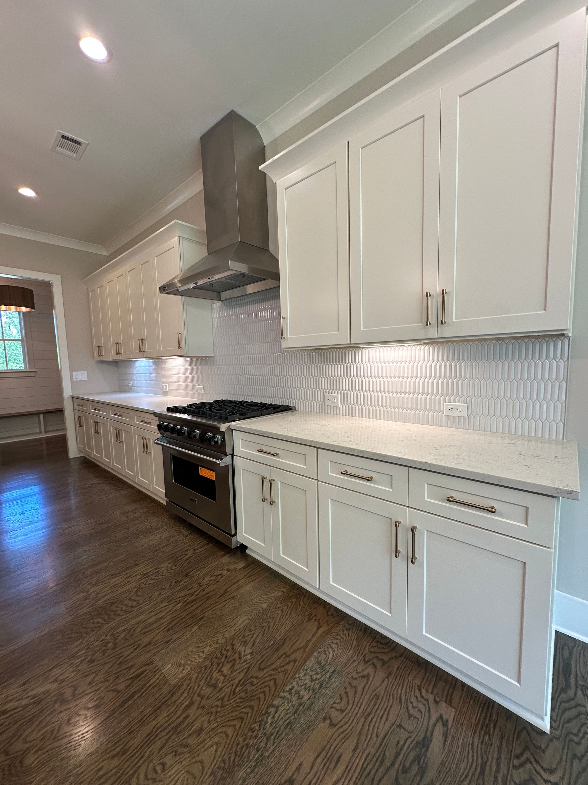 Bright modern kitchen with white cabinets, stainless range hood, and dark wood floor along a long counter.
