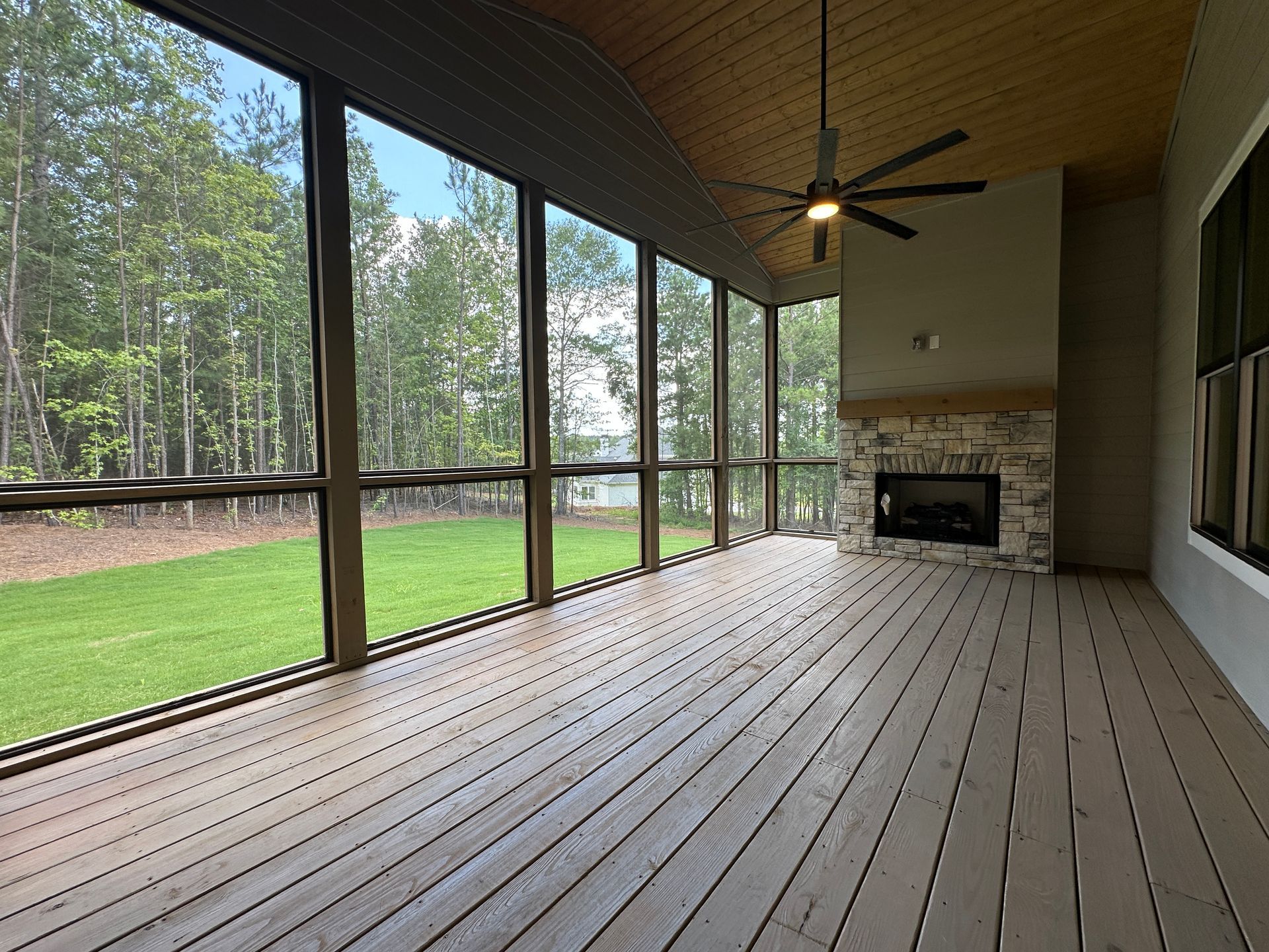 Screened porch with wooden deck, ceiling fan, and fireplace overlooking a green yard and trees.
