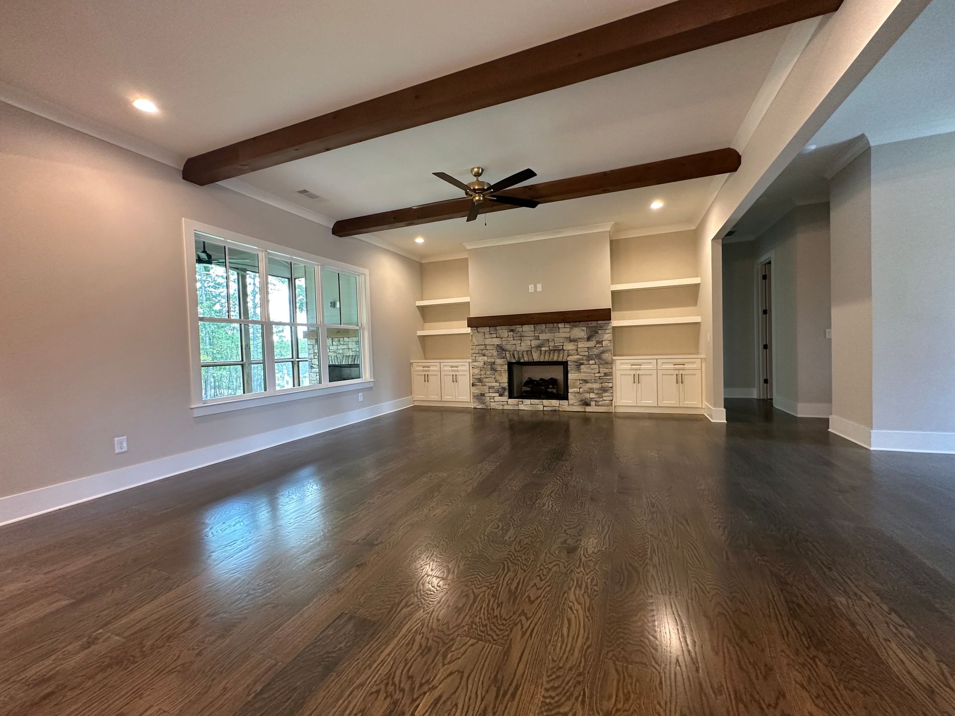 Spacious empty living room with hardwood floors, exposed beams, large window, and stone fireplace