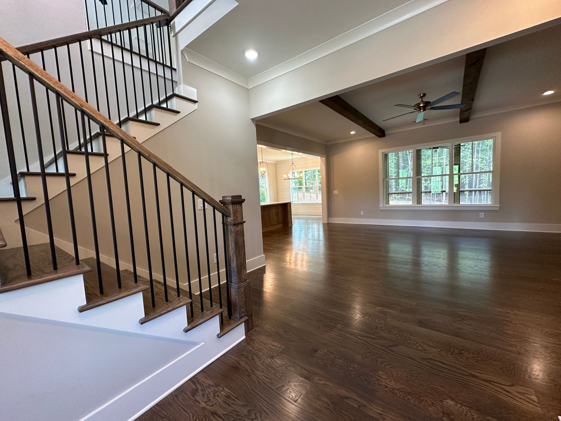 Spacious empty living room with hardwood floors, staircase, and large windows, lit by ceiling lights.