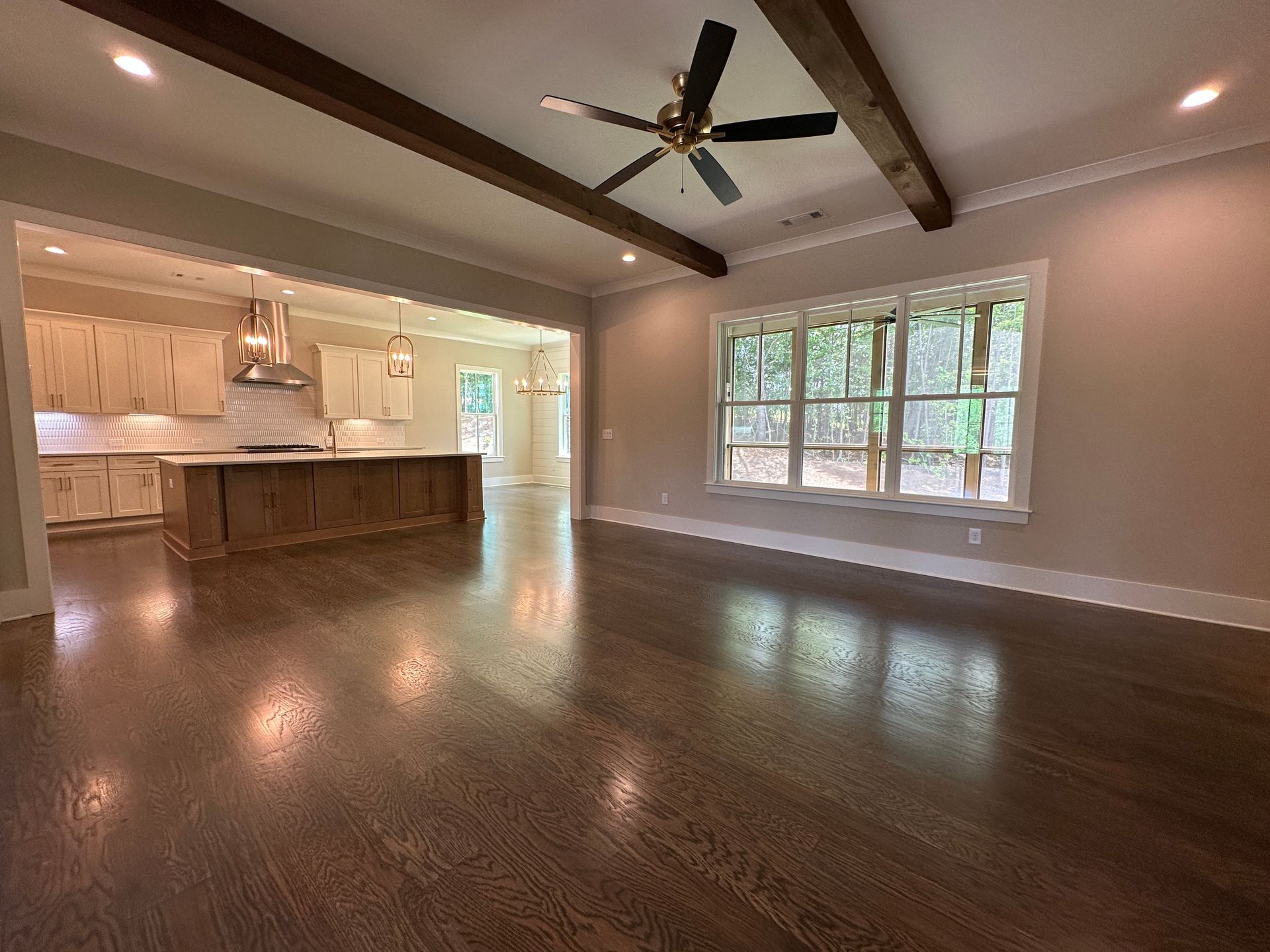 Empty living room with dark wood floors, ceiling fan, and open kitchen with window view