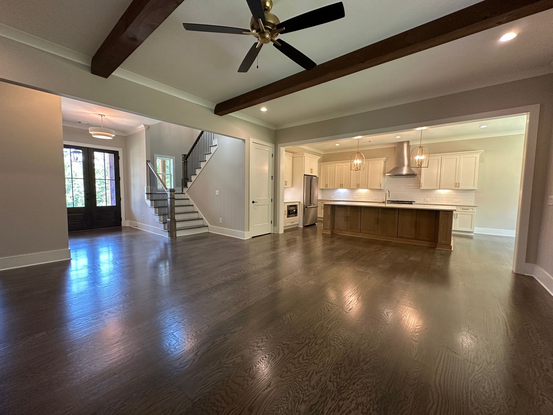 Spacious open-plan living room with dark wood floors, tray ceiling, staircase, and bright kitchen island.