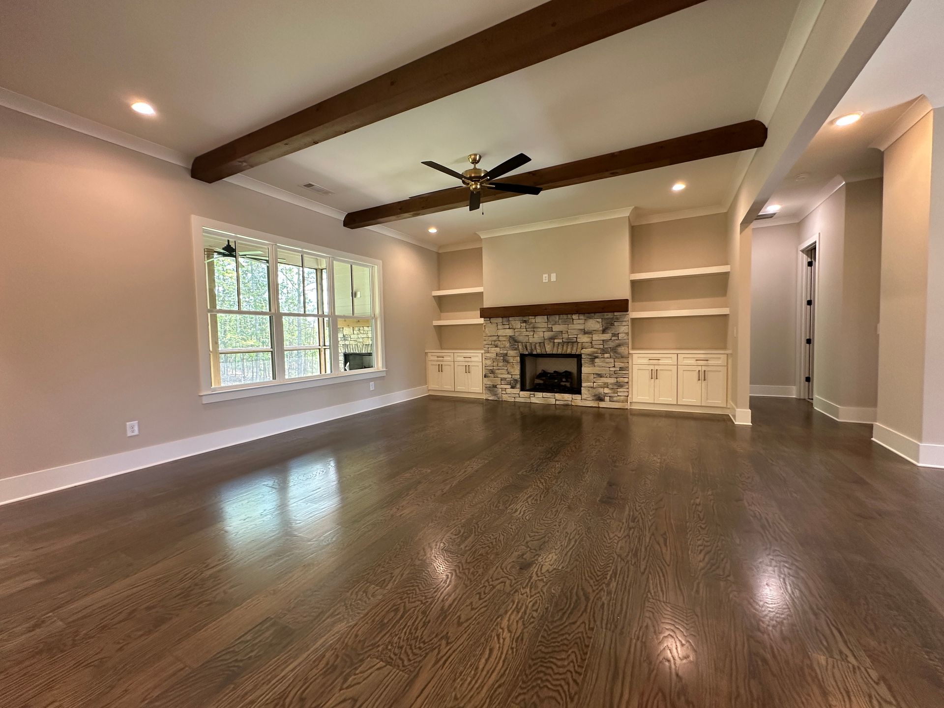 Empty living room with hardwood floors, exposed beams, fireplace, and large window.